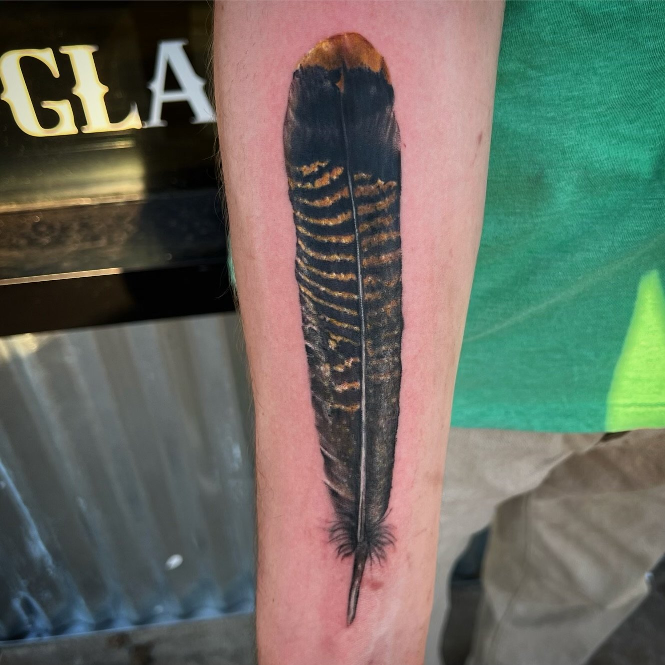 Tattoo of a bird feather on person's forearm with detailed black, brown, and yellow coloring.