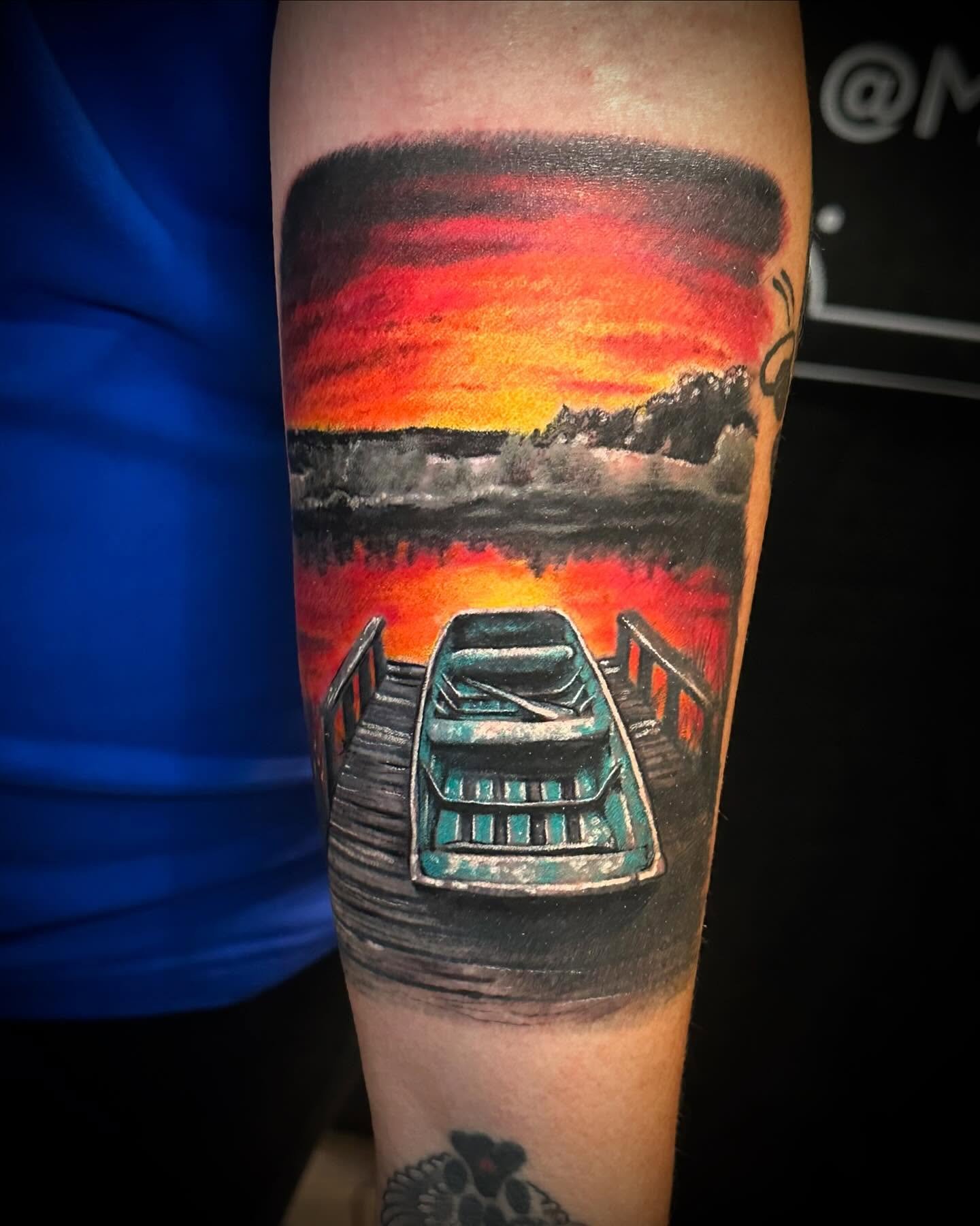 Tattoo of a boat on a lake at sunset with a vivid orange and red sky, black silhouettes of trees, and a dock in the foreground.
