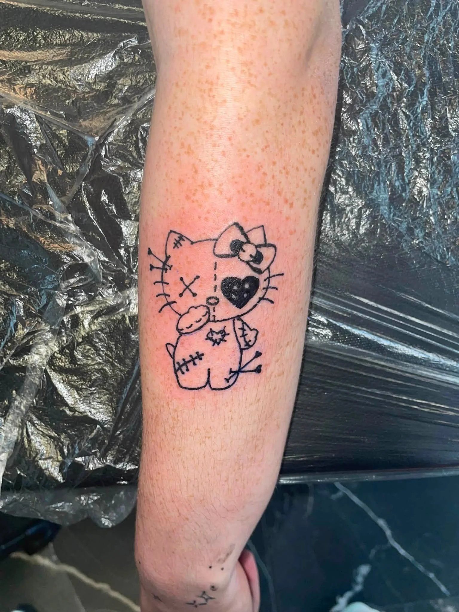 A tattoo of Hello Kitty on a person's forearm, with a black heart, bow, and stitched details, on fair skin with freckles, placed on a black plastic surface.