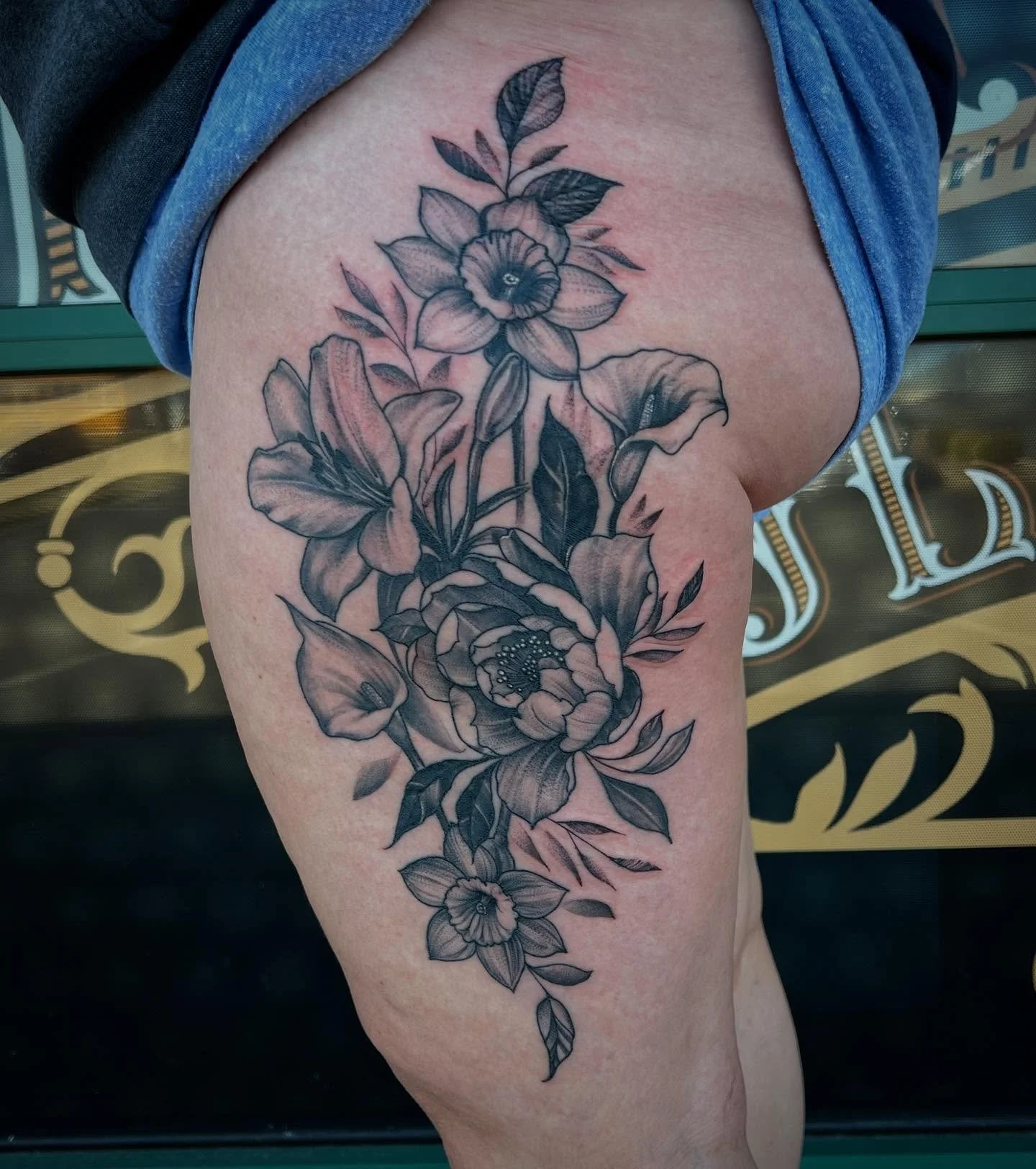 Black and gray floral tattoo on a person's upper thigh, featuring large blooming flowers, leaves, and detailed line work.