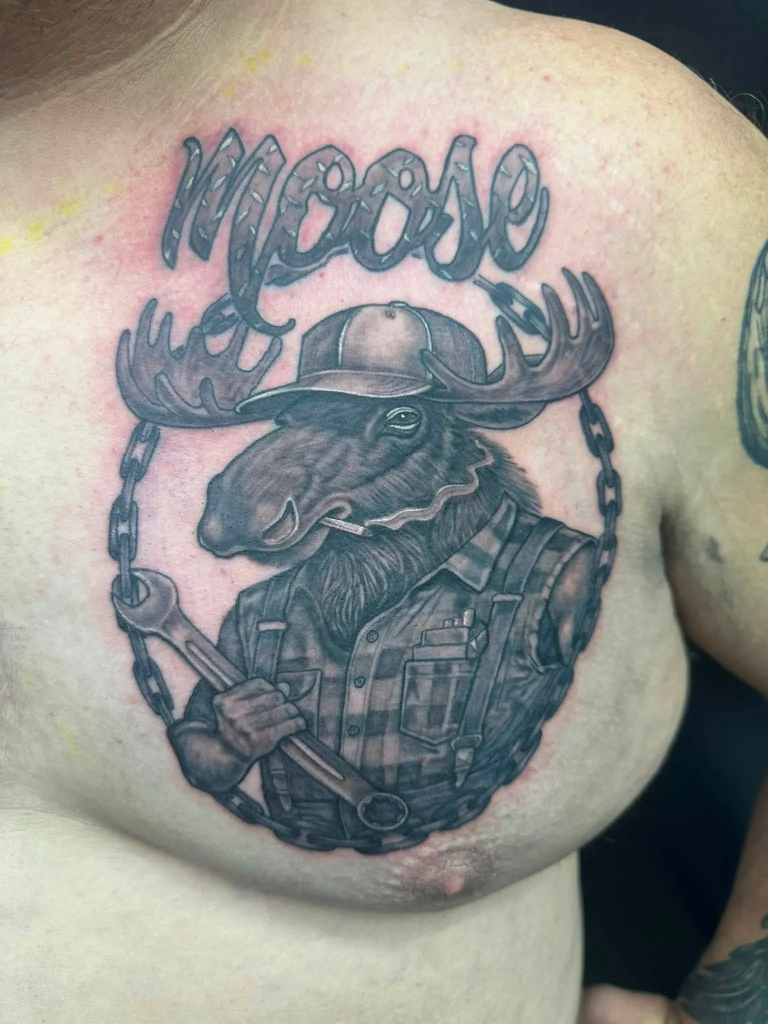 Tattoo of a moose wearing a hat, holding an axe, with chains in the background.