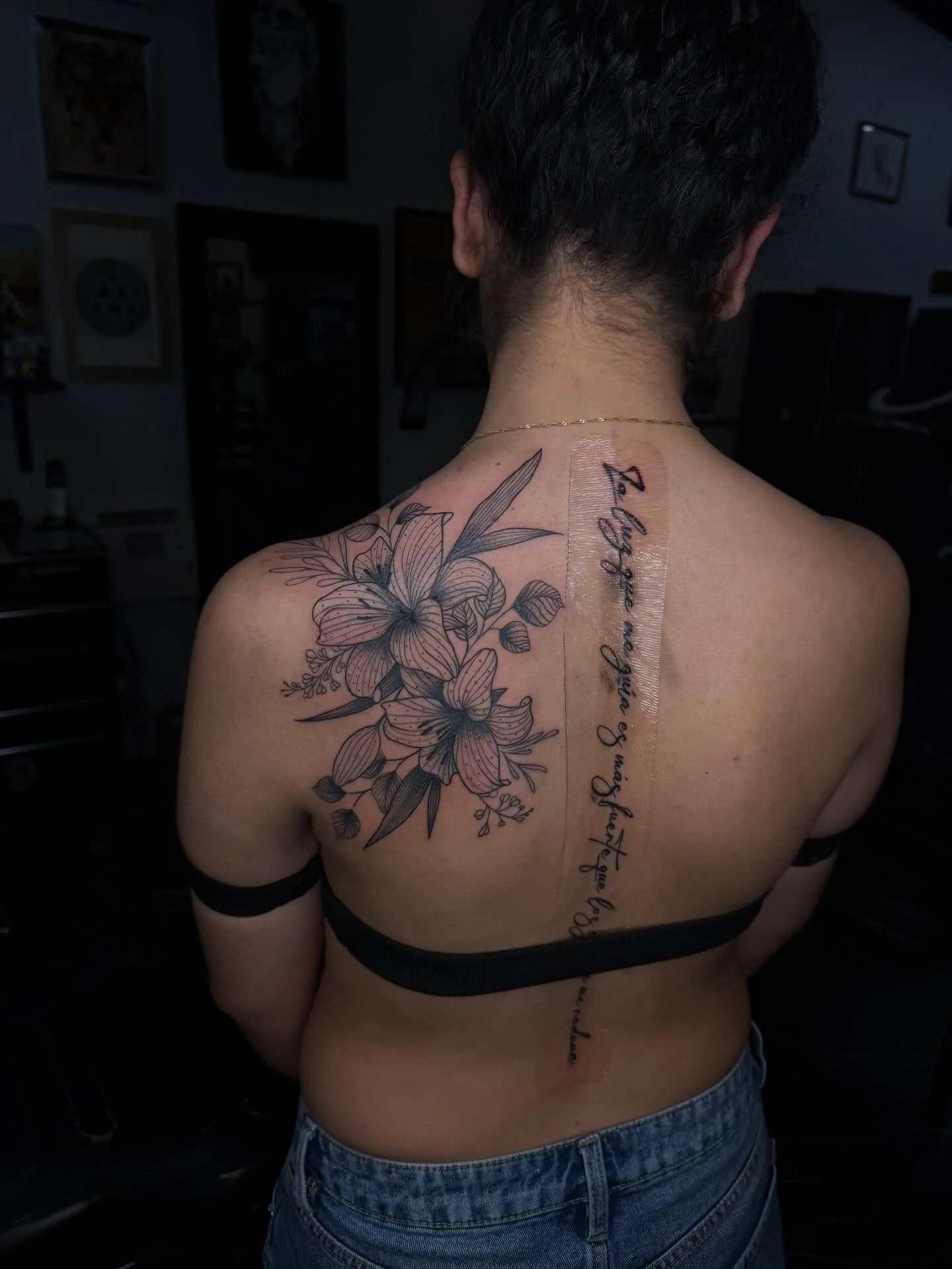 A person with short dark hair has a floral tattoo and cursive writing on their back, with a bandage covering part of the tattoo.