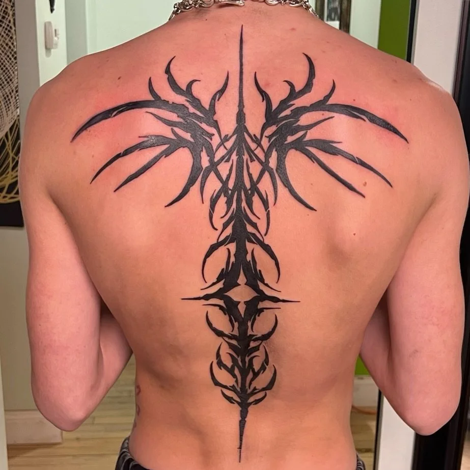A person with a large tribal-style black tattoo on their back, extending from the upper back down to the lower back, with symmetrical, sharp, curved lines creating an intricate design.
