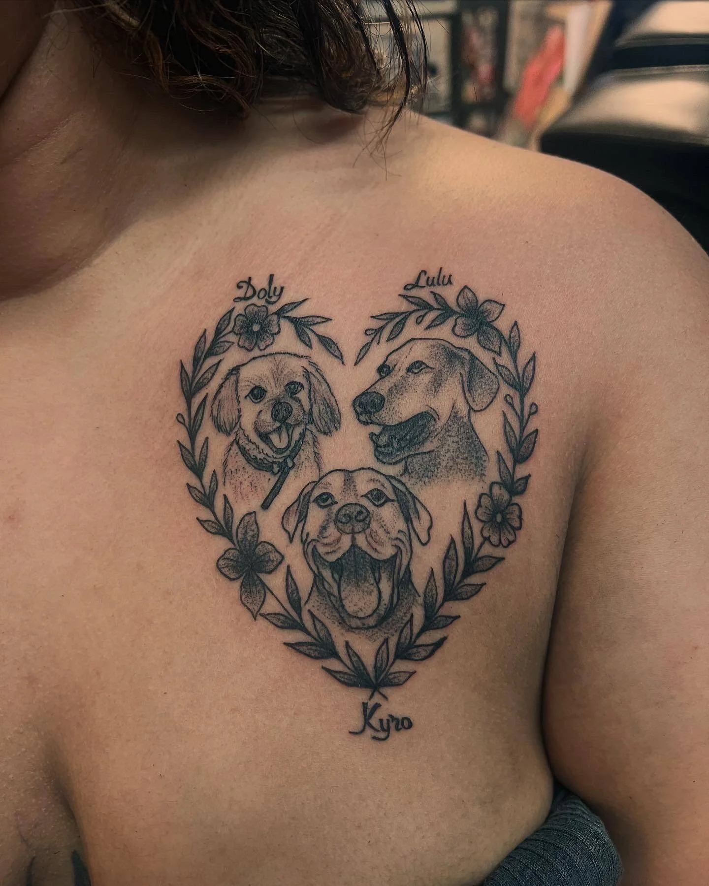 A tattoo of three dogs surrounded by a floral wreath, with the names "Doly," "Lulu," and "Kyro" written above each illustration.