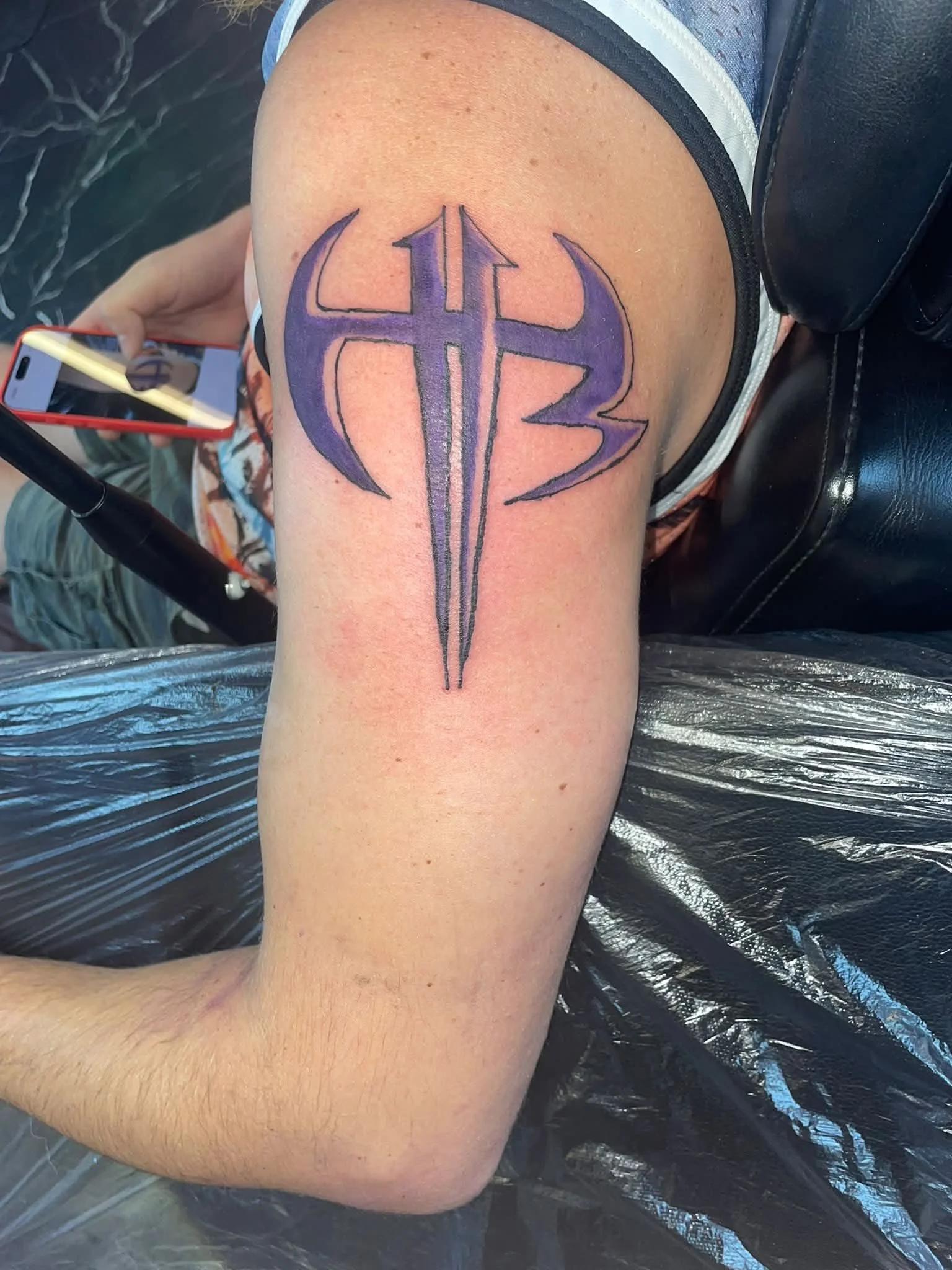 A fresh tattoo on a person's upper arm featuring a stylized purple symbol with black outlines, placed over slightly reddened skin.