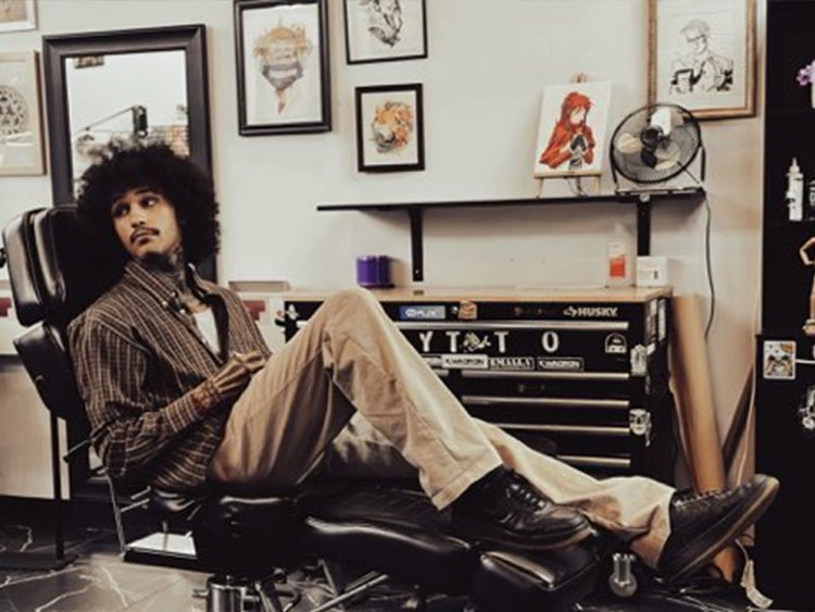A person with curly hair is sitting on a chair in a tattoo studio surrounded by artwork and tattoo equipment.