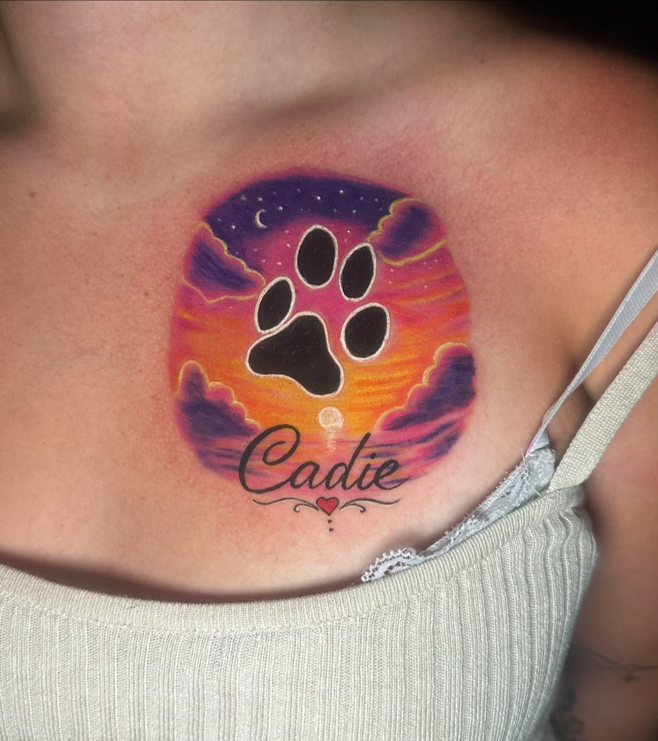 Tattoo on skin featuring a sunset with purple clouds, a black paw print in the center, and the name 'Cadie' in cursive with a small red heart underneath.