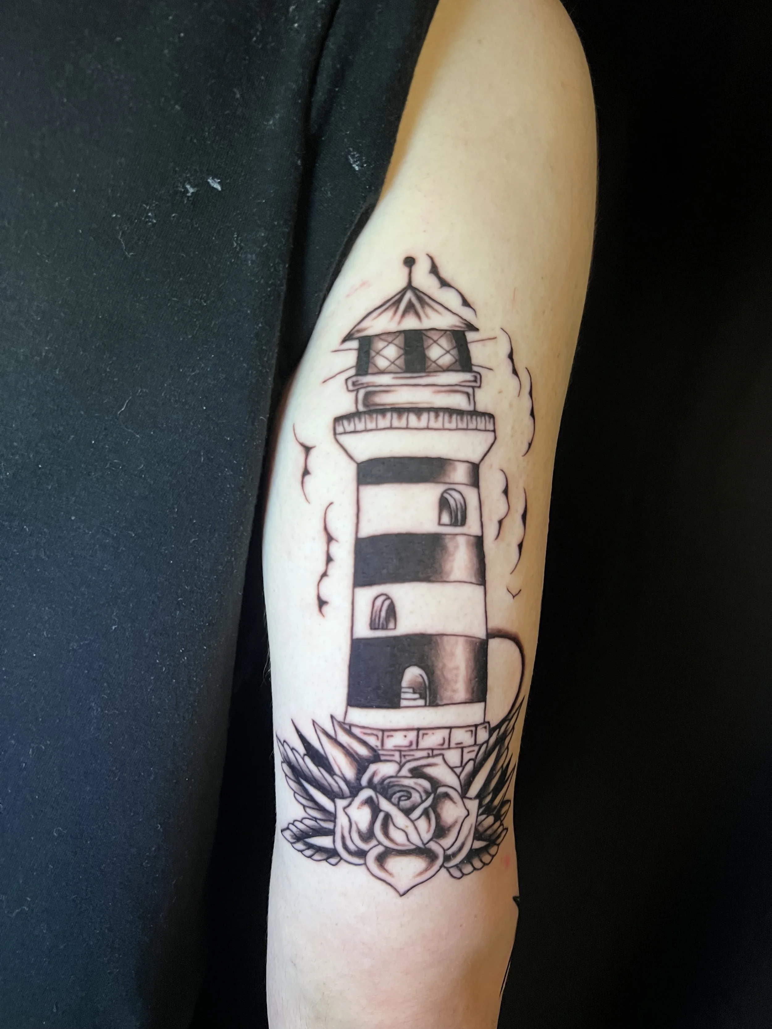 Black and white tattoo of a lighthouse surrounded by waves, clouds, and intricate decorative elements on a person's arm.