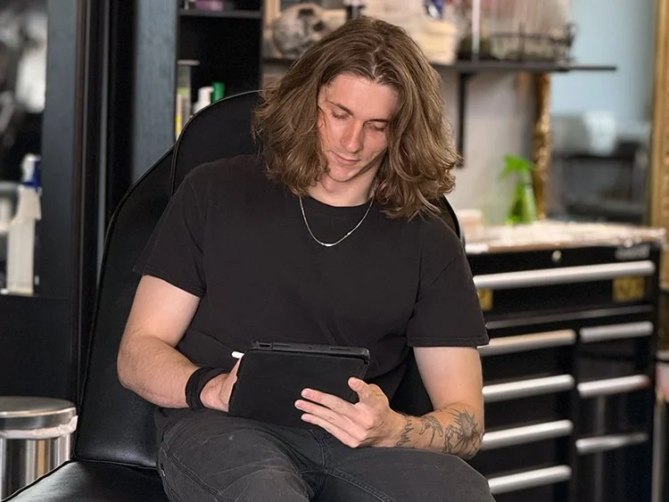 Young person with long brown hair, wearing a black t-shirt, sitting on a black chair in a workshop or studio, using a tablet.