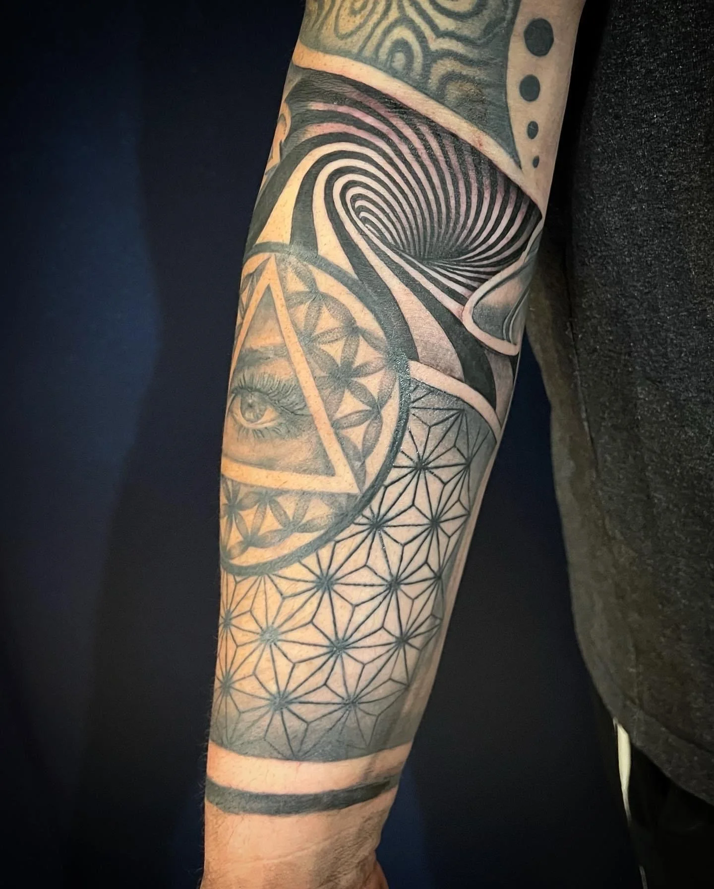A person's arm with multiple black ink tattoos, including a geometric pattern, an eye inside a triangle, and a large hypnotic whirlpool design.