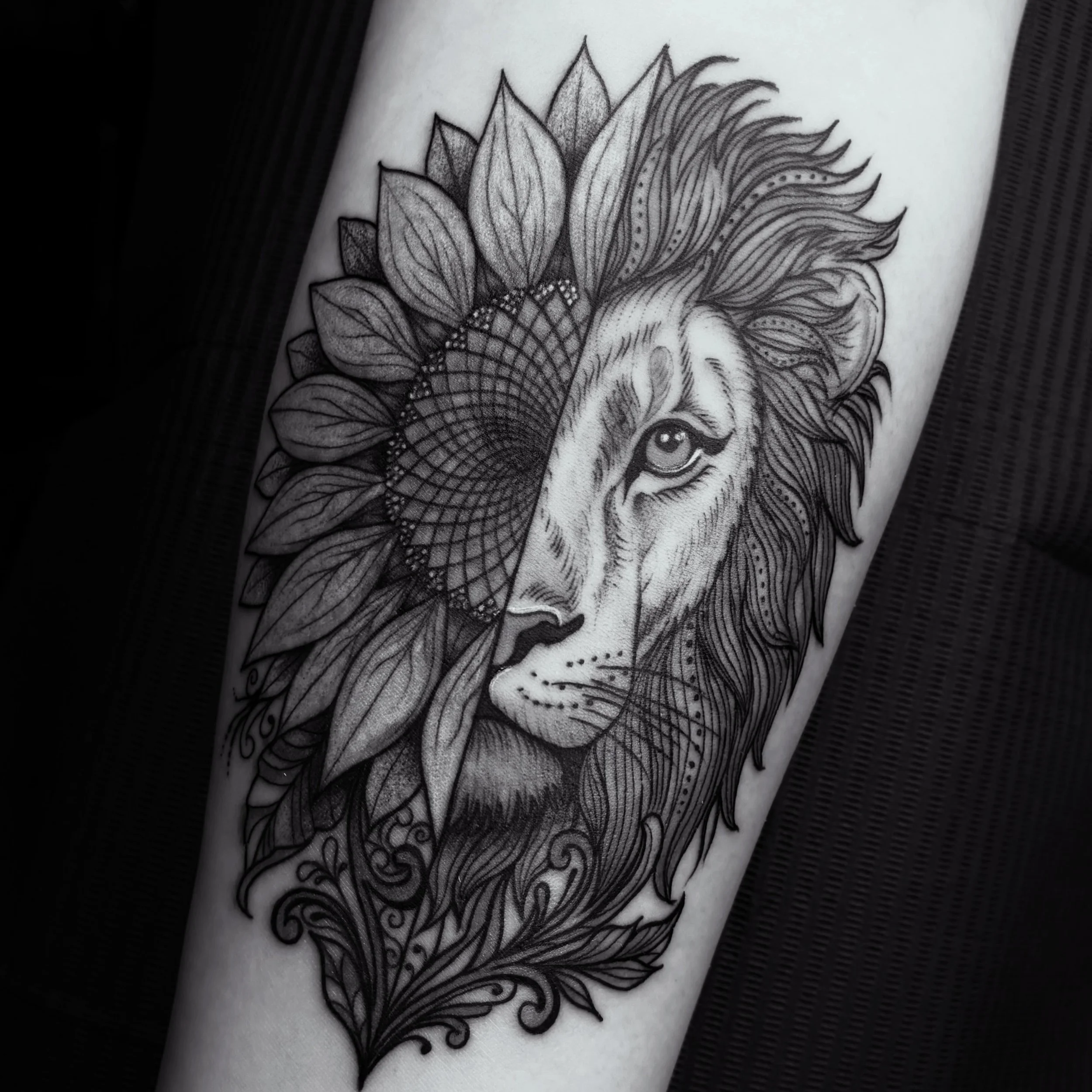 Black and gray tattoo of a lion with a mane, surrounded by detailed floral and geometric patterns.
