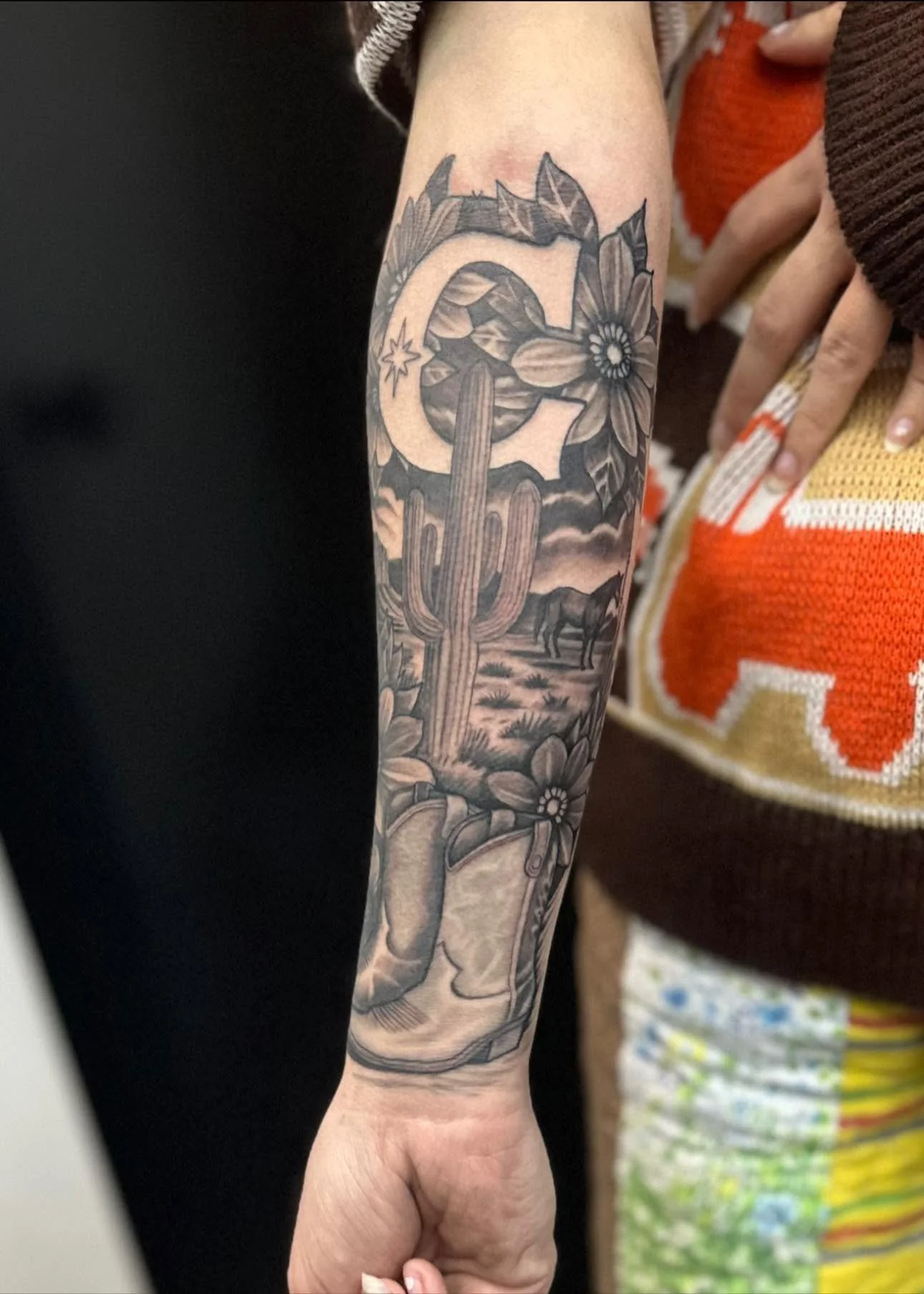 A person's arm with a detailed black and gray tattoo featuring a large cactus, a flower, and a landscape with a horse in the background.