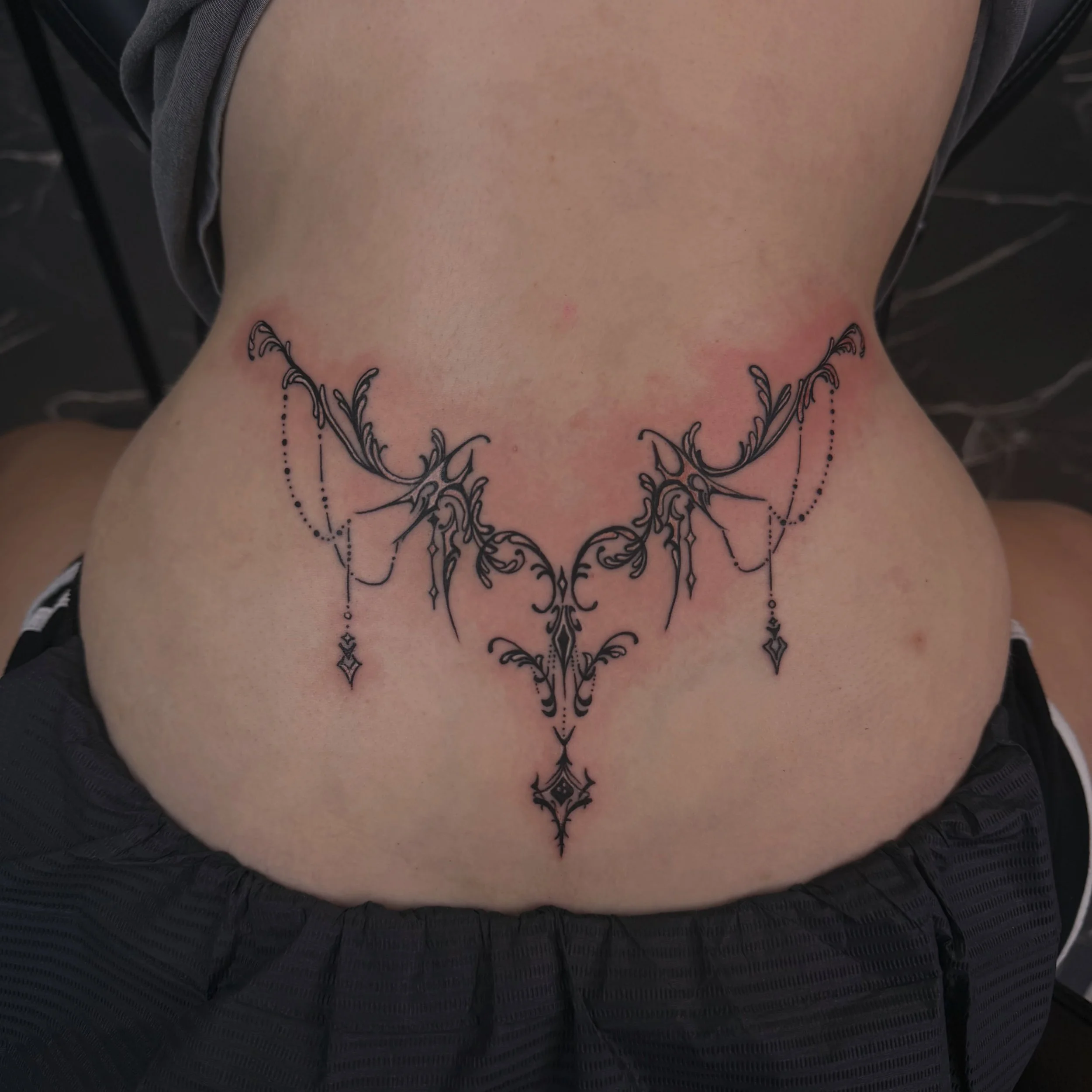 Close-up of a person's lower back and waist area, showing a black ornamental tattoo with symmetrical floral and swirl patterns.
