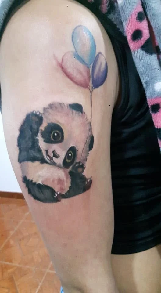 Tattoo of a cute panda holding two balloons, one blue and one purple, on a person's upper arm.