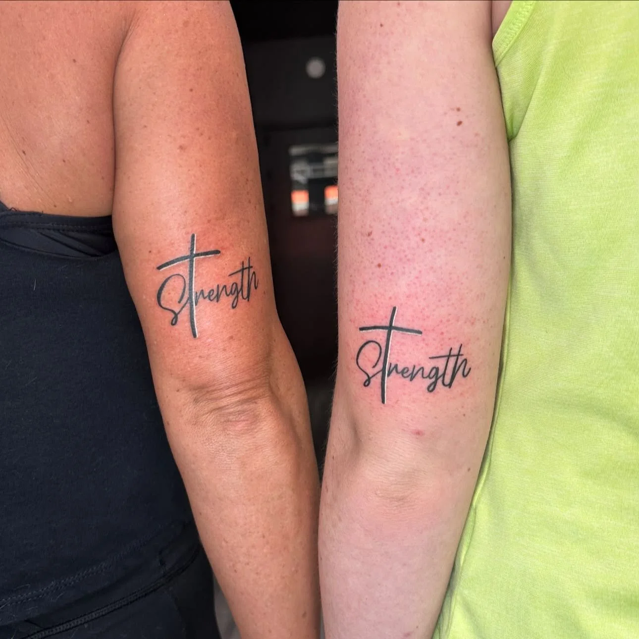 Two people with matching tattoos that say 'Strength' on their upper arms. The tattoos feature a cursive and cross design.