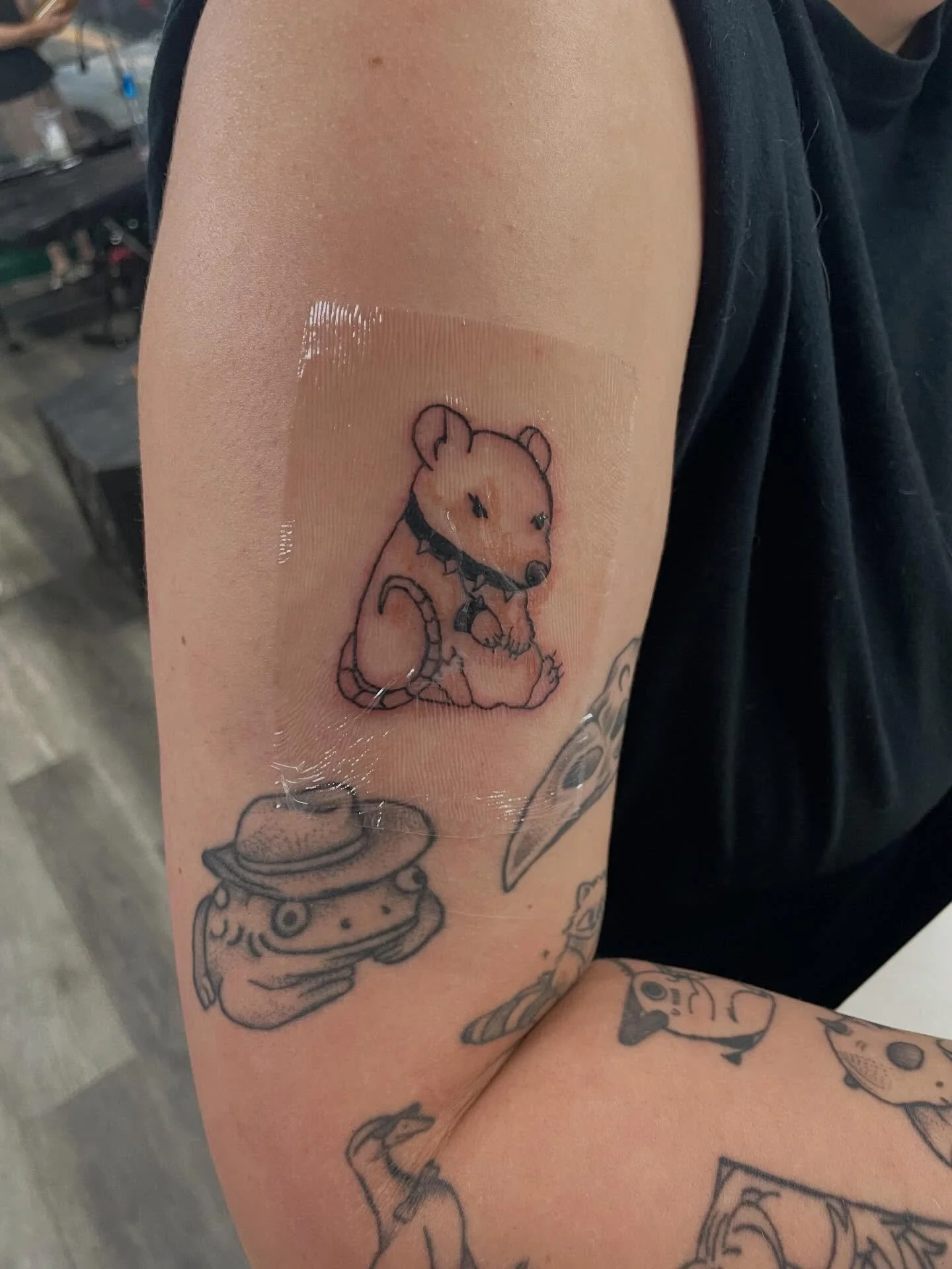 A person's upper arm with several tattoos, including a cartoon-style dog wearing a spiked collar, and other tattoos of a burger and various images.