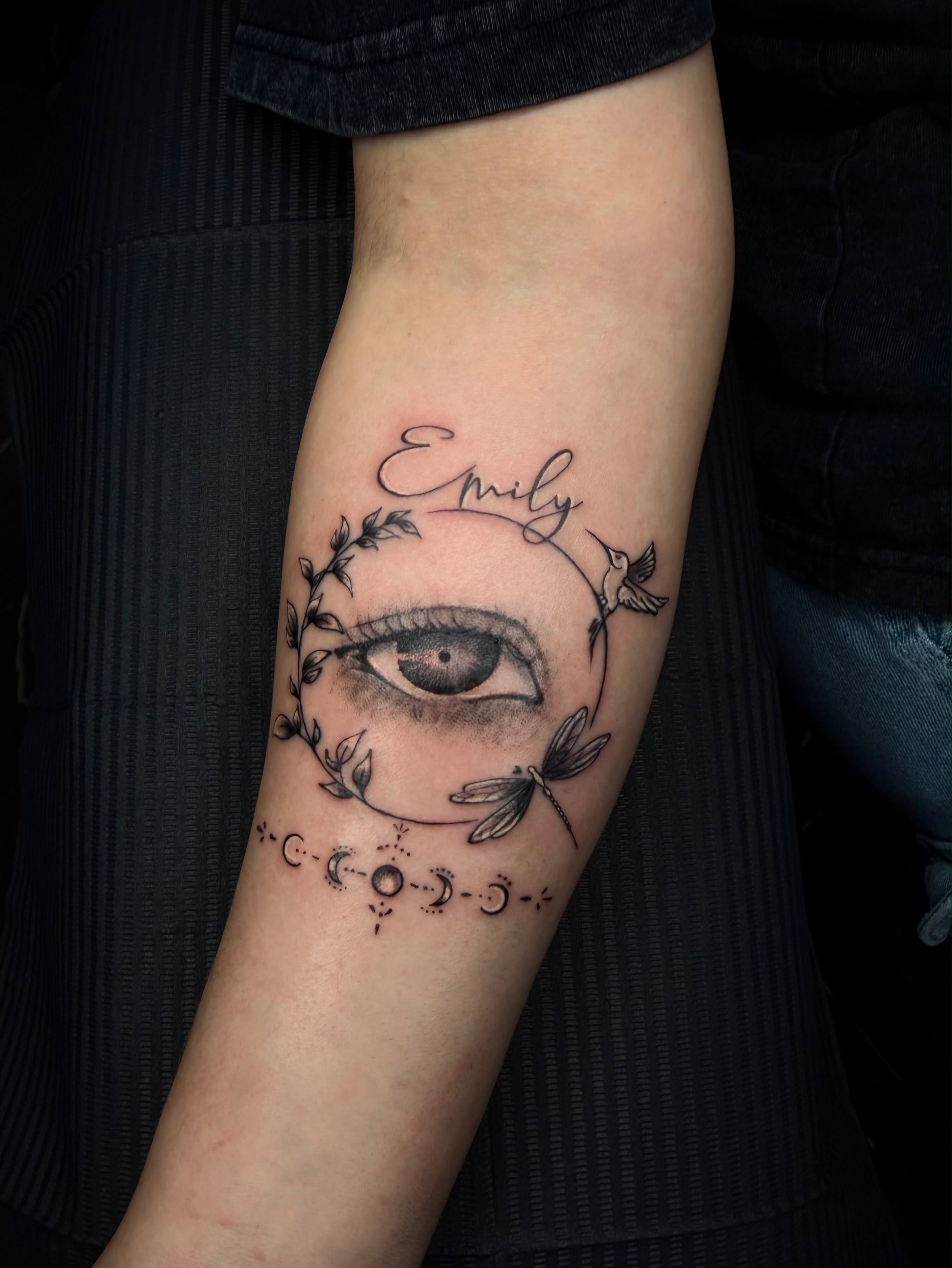 A person's arm with a tattoo of the word 'Emily' in cursive above an eye with decorative leaves and a hummingbird, and a moon phase and celestial symbols at the wrist.