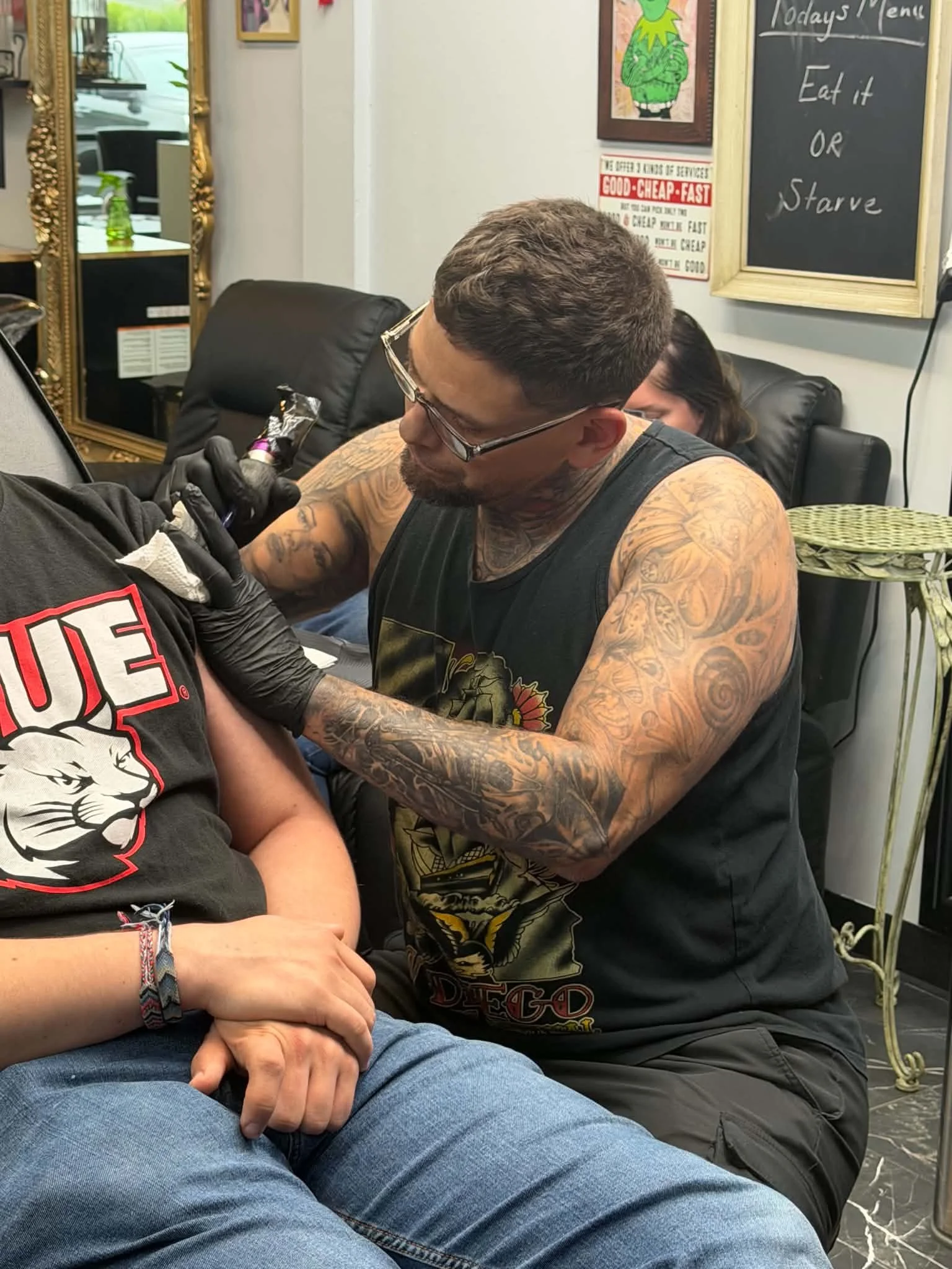 A man with tattoos on his arms and glasses is getting a tattoo on his arm. There is a person with a black glove tattooing him and another person sitting behind him. The setting appears to be a tattoo studio with chairs, a mirror, and wall art.