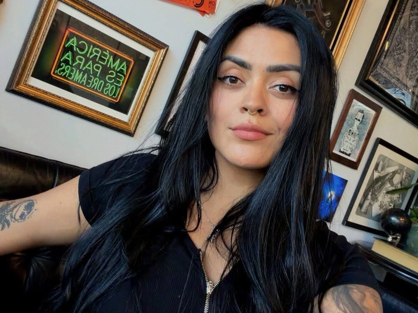 A woman with long black hair and tattoos on her arm, sitting in a restaurant or cafe, with framed artworks and a neon sign in the background.
