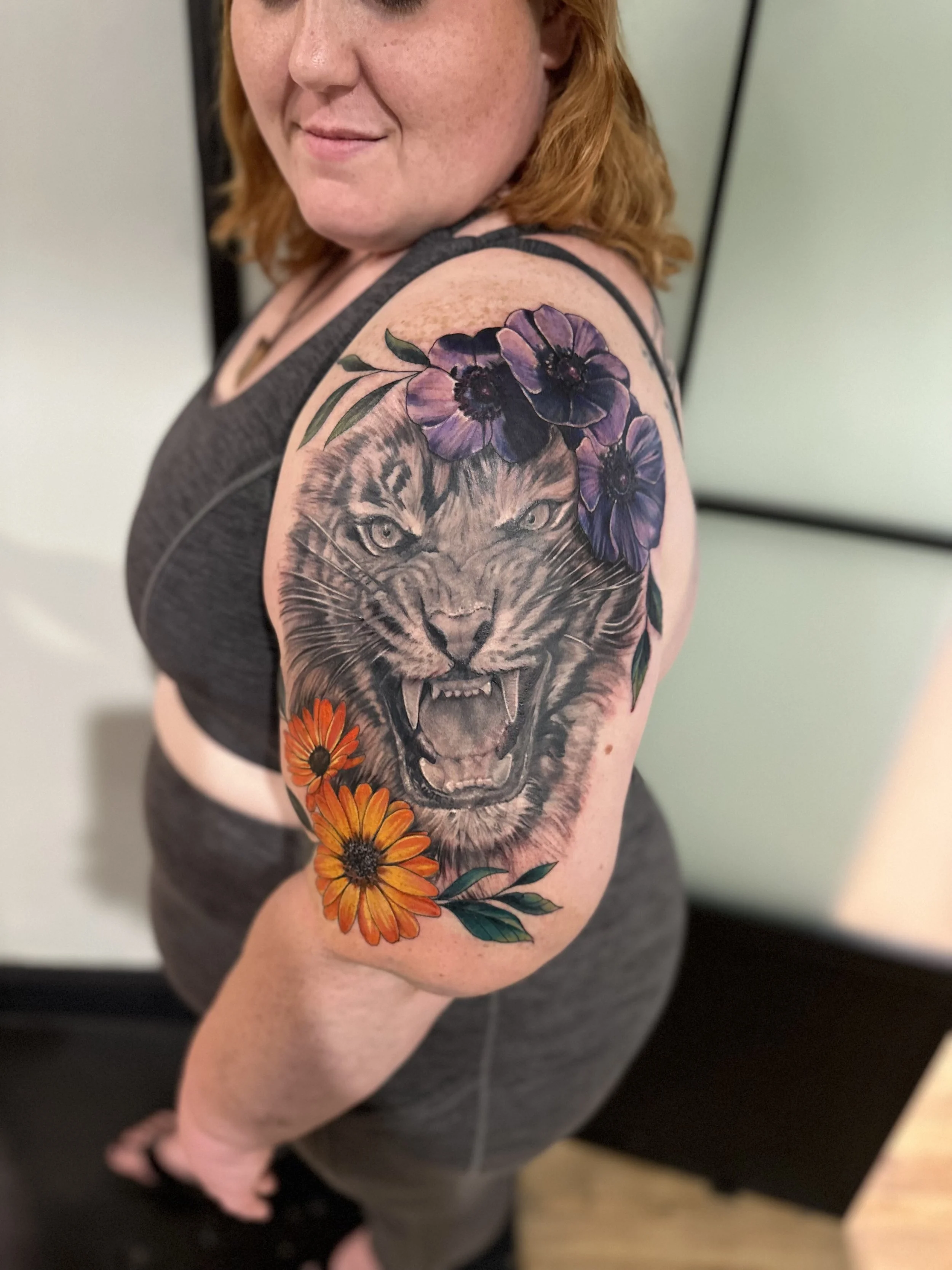 A woman with red hair and freckles showing her tattooed upper arm featuring a detailed lion's face intertwined with purple flowers and orange daisies.