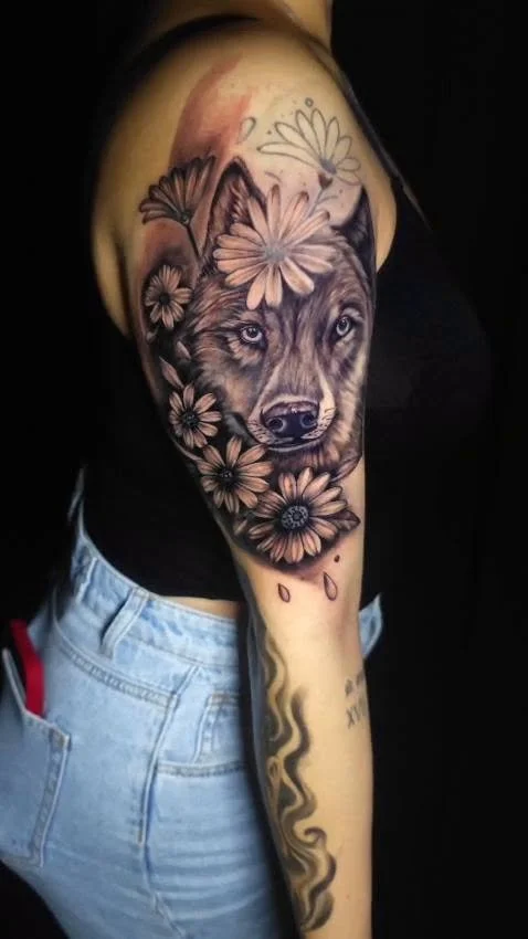 Tattoo of a wolf's face surrounded by daisies and other flowers on a person's upper arm.