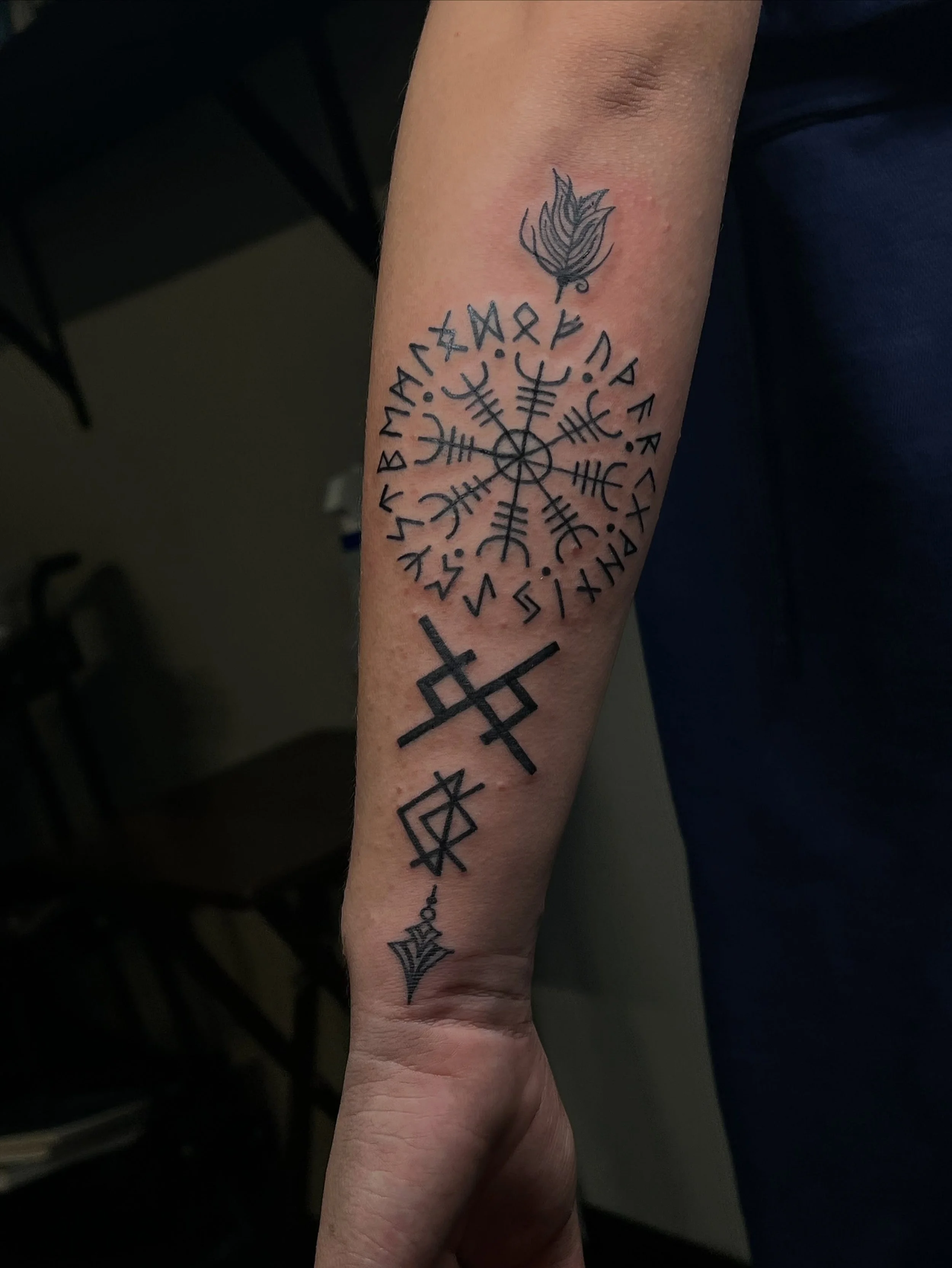 Tattoo of a flame at the top of the forearm with a circular geometric symbol and runic inscriptions below.