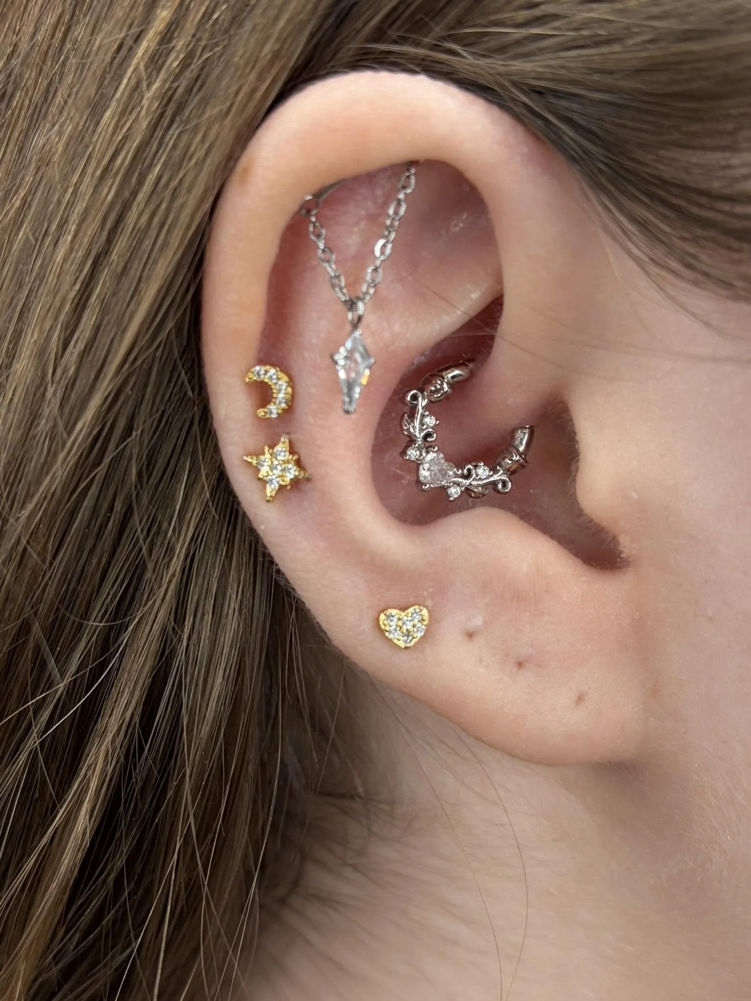 Close-up of a person's ear with multiple earrings, including a gold crescent moon, a gold star, a silver chain with a pendant, a silver ring, and a heart-shaped earring.