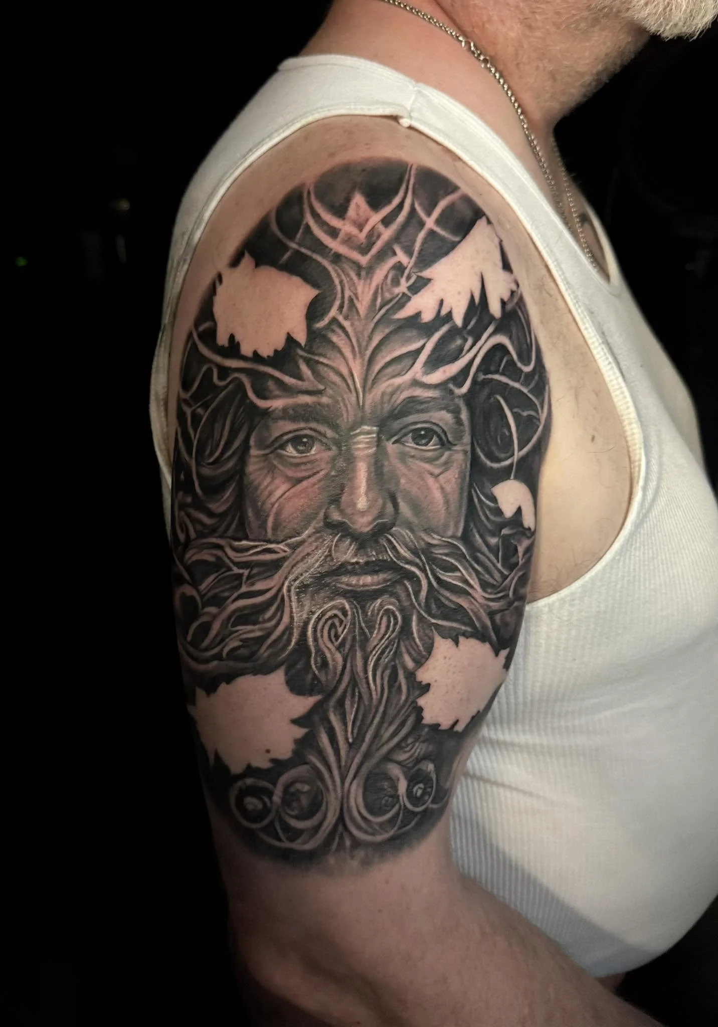 A tattoo of a face on a person's upper arm, with detailed features and intricate linework surrounding the face, resembling a bearded man with a crown of clouds or flames.