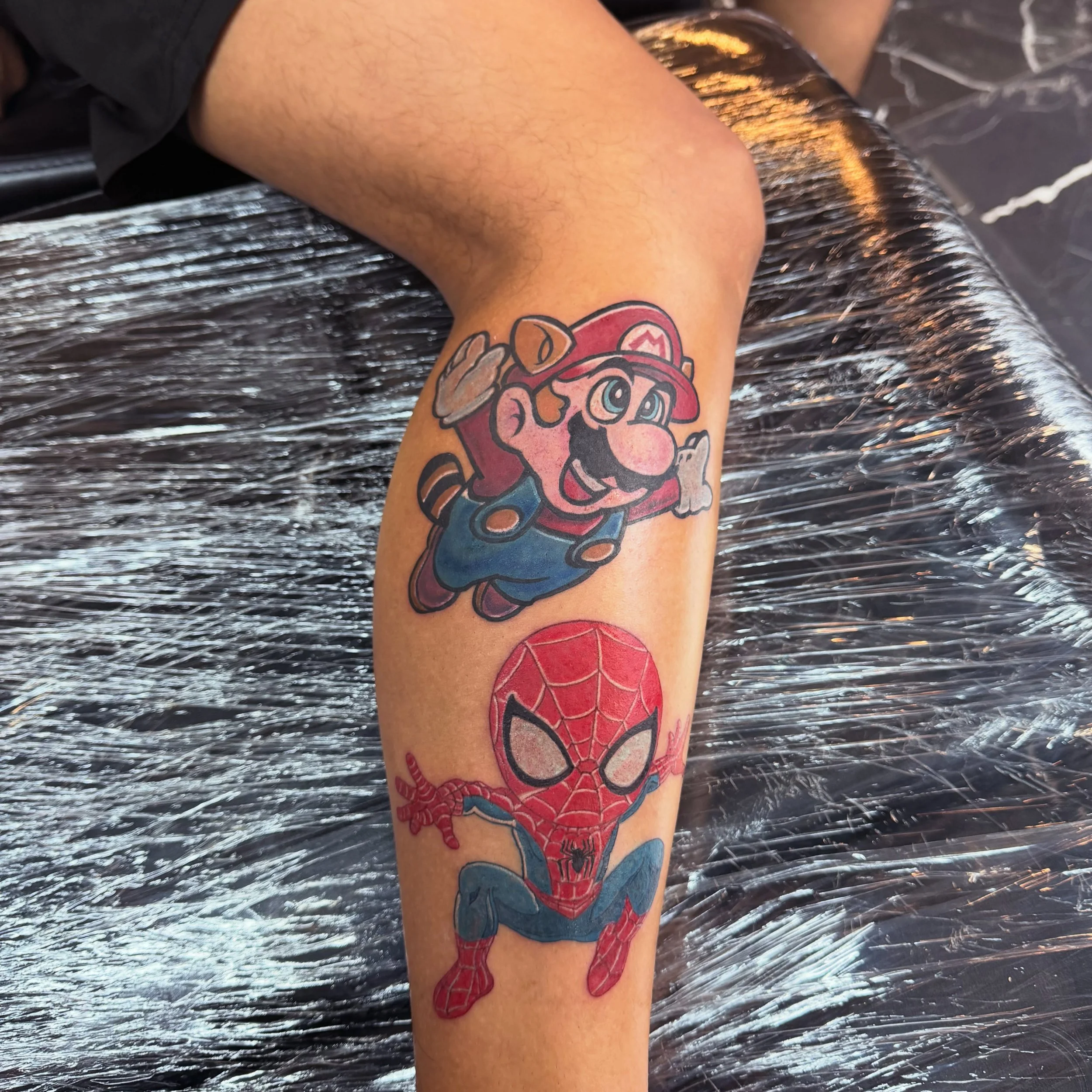 A person's arm with two tattoos: one of Mario wearing his signature red hat, and another of Spider-Man's comic-style face.