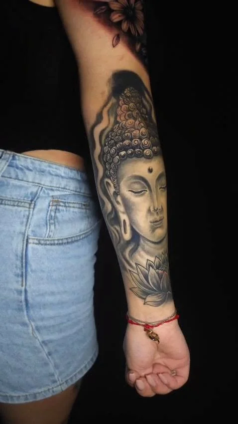 Tattoo of Buddha's face with closed eyes, curly hair, and a lotus flower at the wrist.