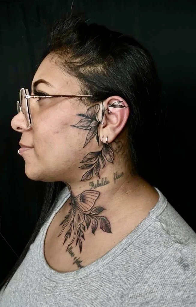 Profile view of a woman with black hair, wearing glasses, with multiple tattoos including a butterfly on her neck, leaves around her ear, and text tattooed on her neck.