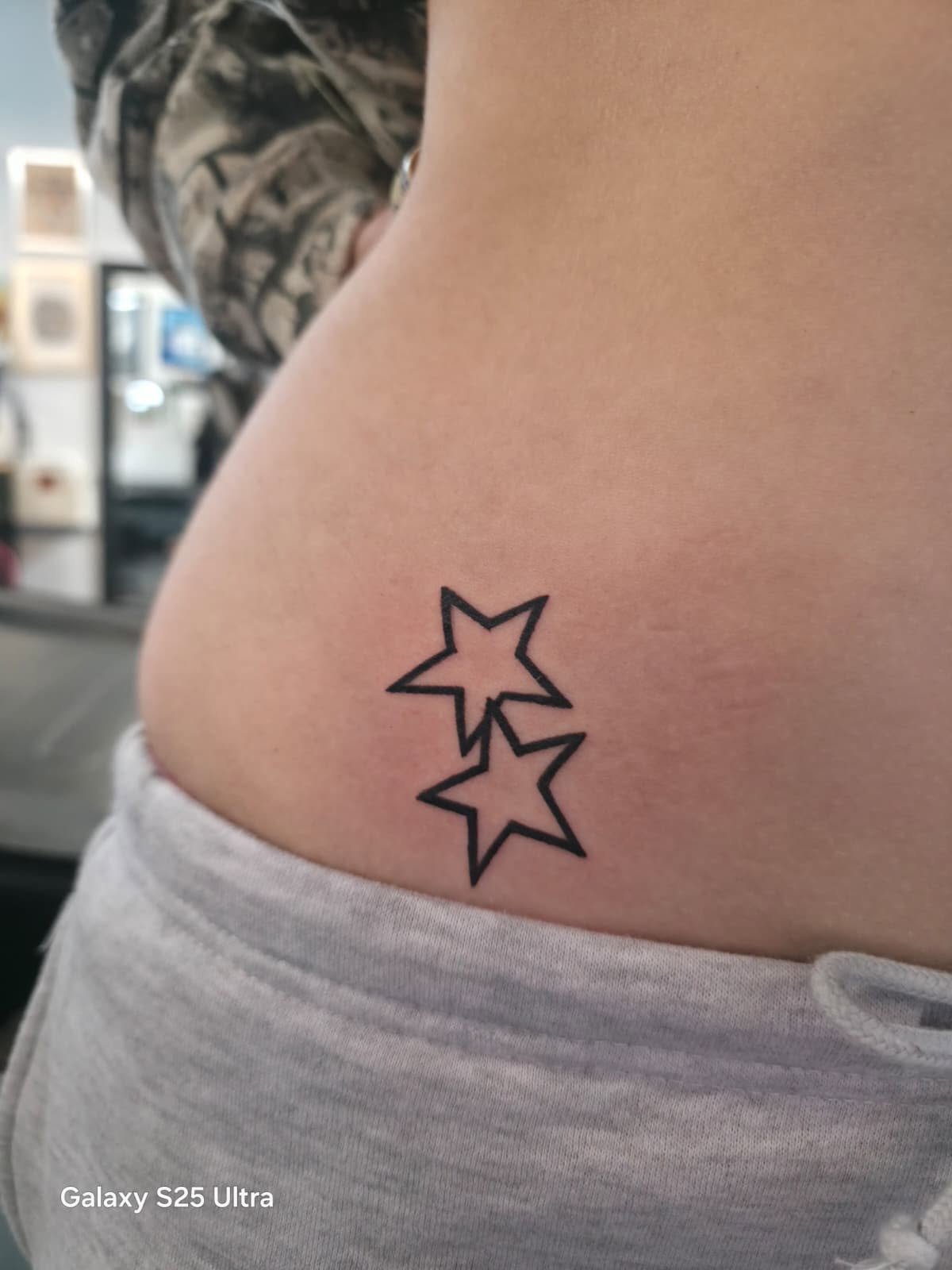 Tattoo of two outlined stars on person's side, with the person wearing a gray shirt and a camouflage sleeve visible in the background.