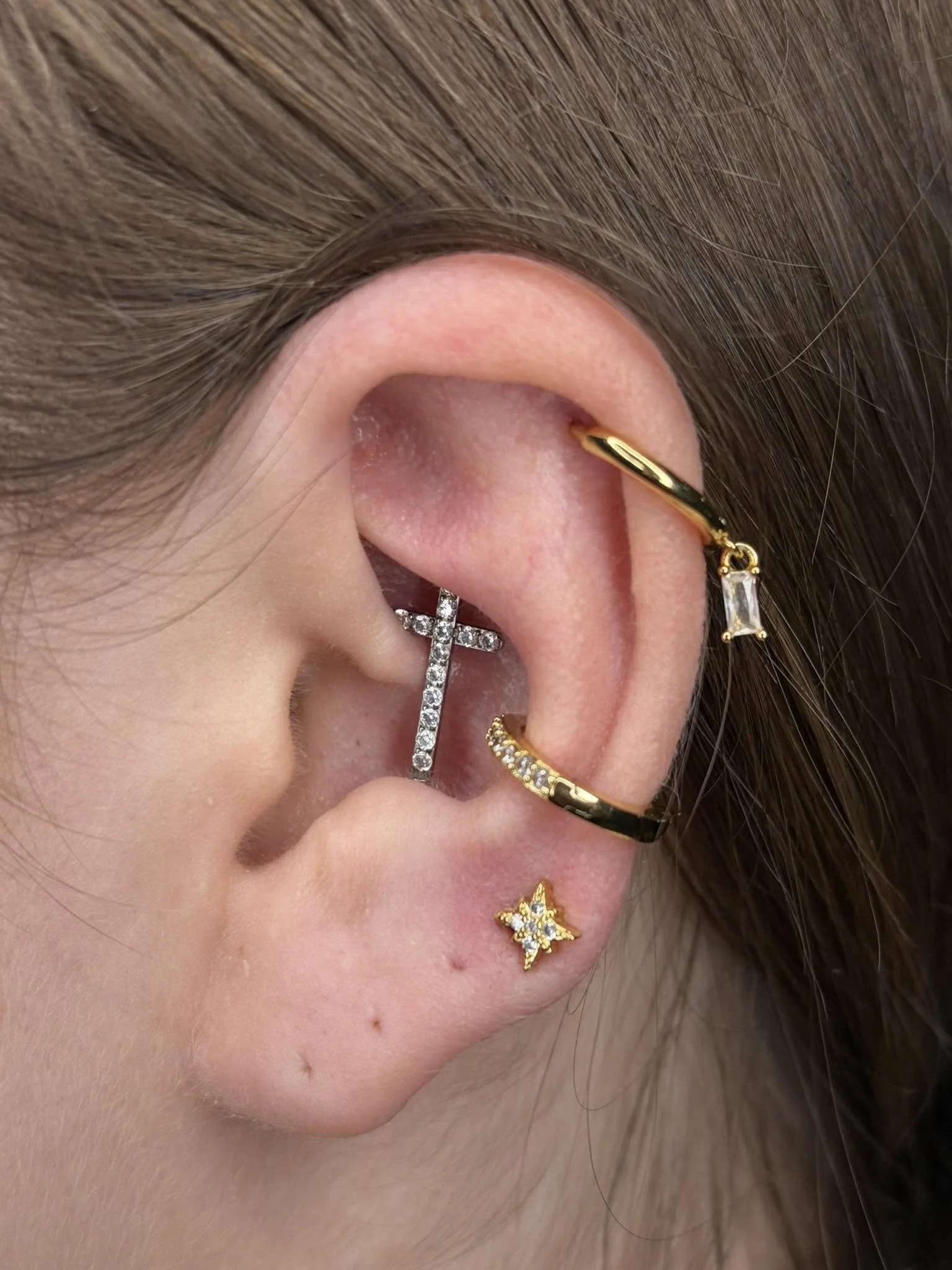 Close-up of a person's ear adorned with multiple earrings, including a gold hoop with a rectangular gemstone, a silver cross-shaped earring with rhinestones, and a star-shaped earring with rhinestones, along with brown hair.