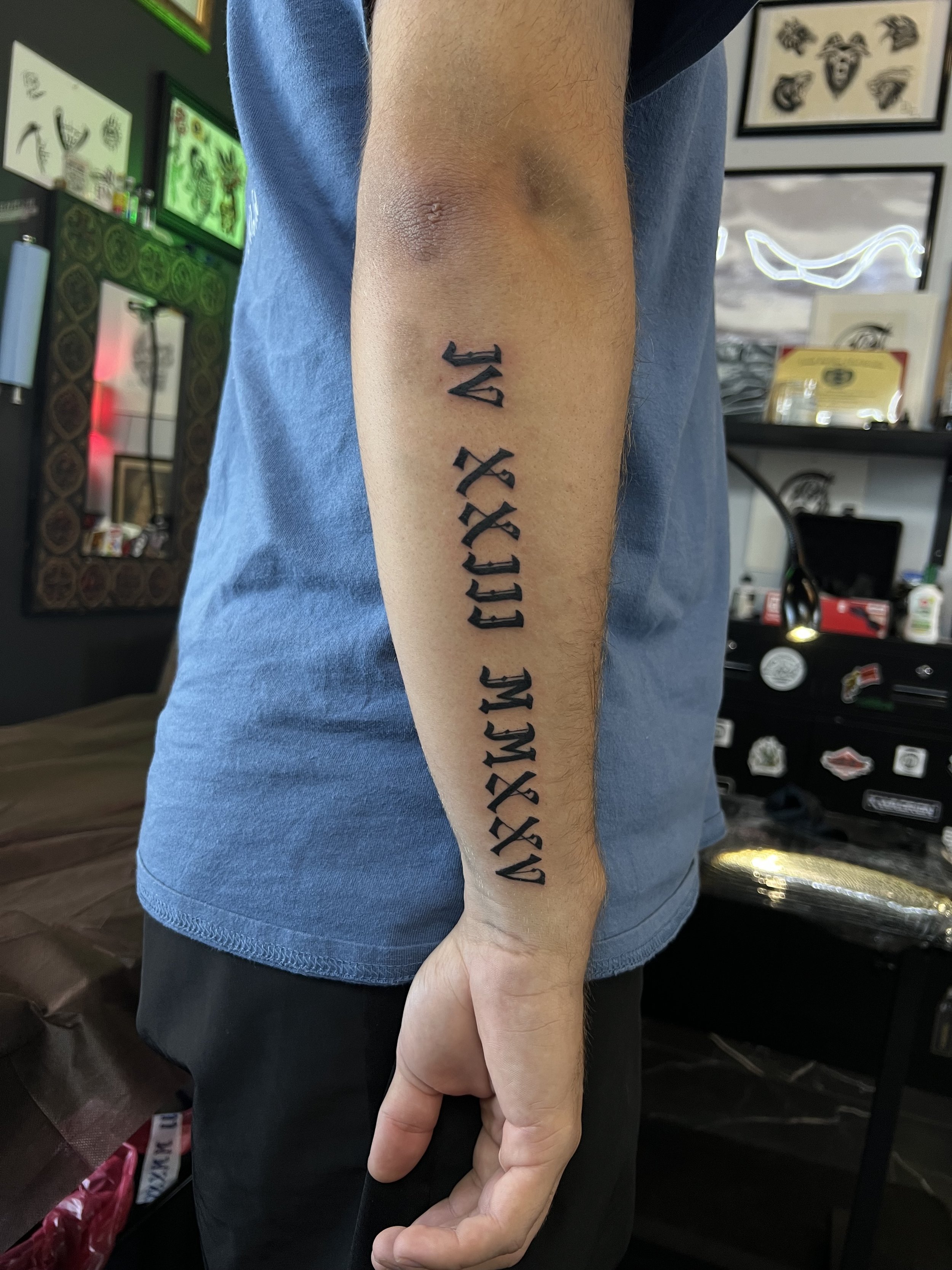 A person's arm with a tattoo of black Gothic-style text, with a background of tattoo art and decorations in a tattoo shop.