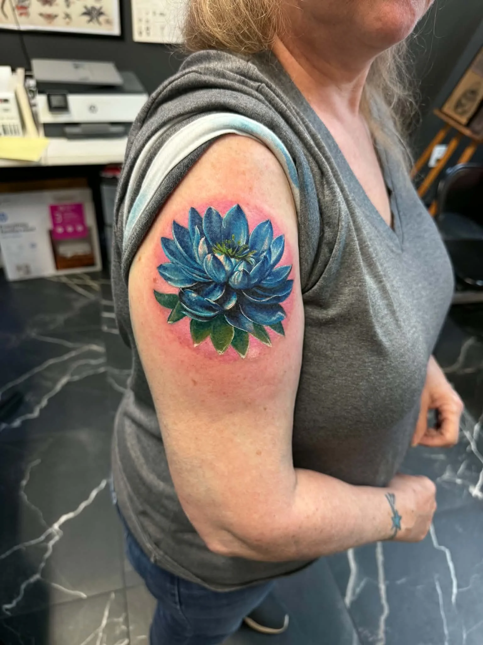 A woman with a large blue and green succulent flower tattoo on her upper arm, wearing a gray sleeveless shirt.