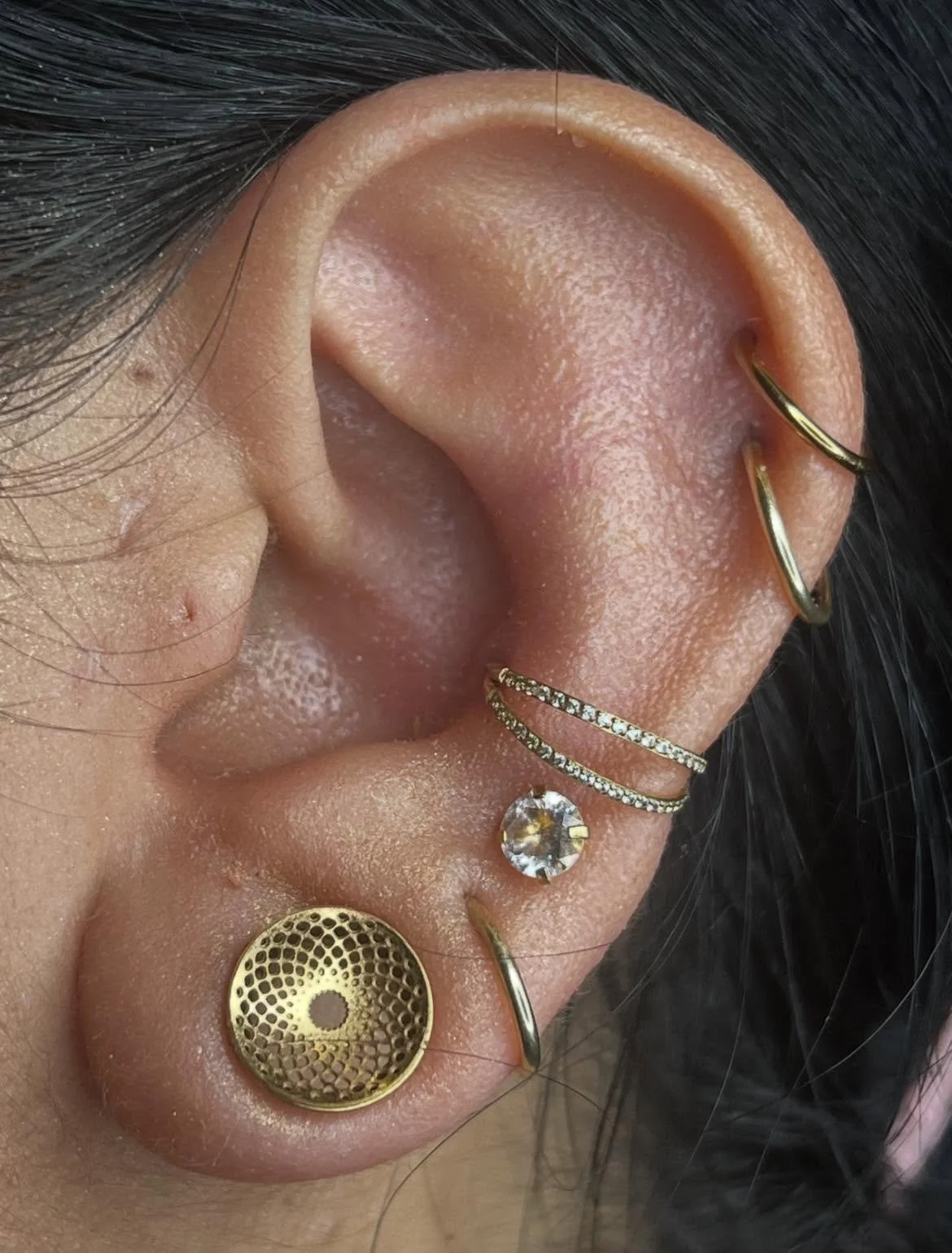 Close-up of a person's ear with multiple piercings, including a gold hoop in the upper cartilage, a diamond-studded hoop, and a large diamond stud in the lower lobe, along with gold earrings featuring intricate designs.