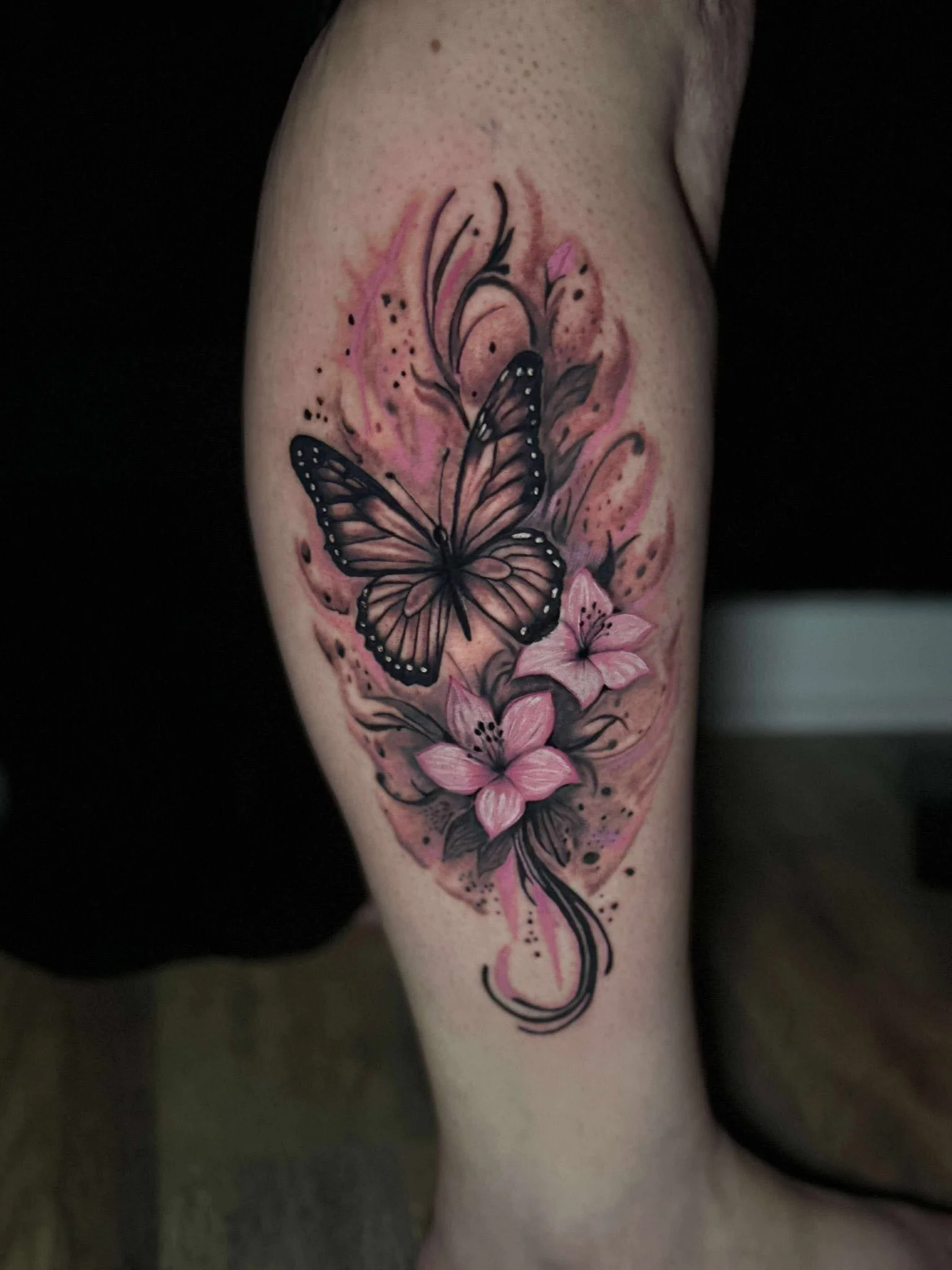 Tattoo of a butterfly with black and pink accents on a person's arm, surrounded by pink flowers and watercolor-style splashes.