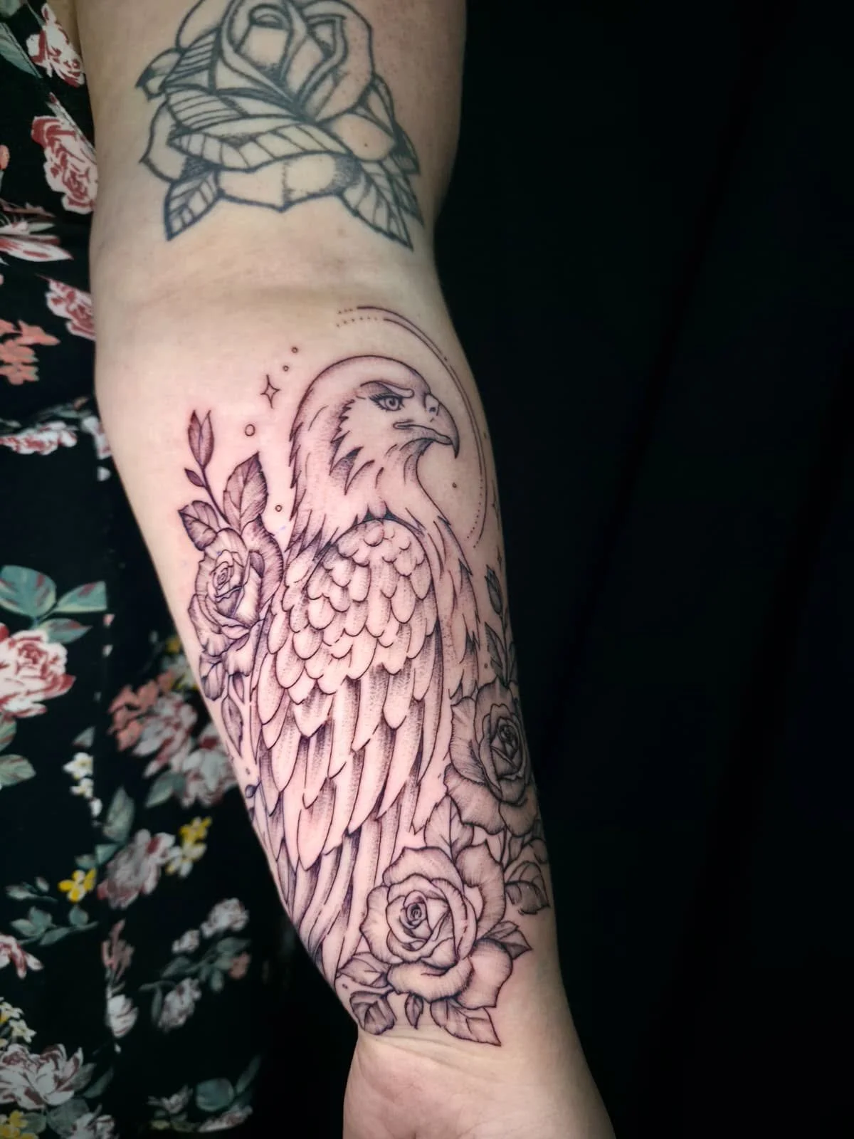 A black and gray tattoo of an eagle with roses and leaves on a person's arm.