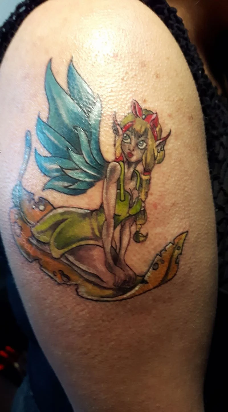 Colorful fairy tattoo on arm featuring a fairy with blue wings, blonde hair, elf-like ears, and wearing a green dress, sitting on a pumpkin.