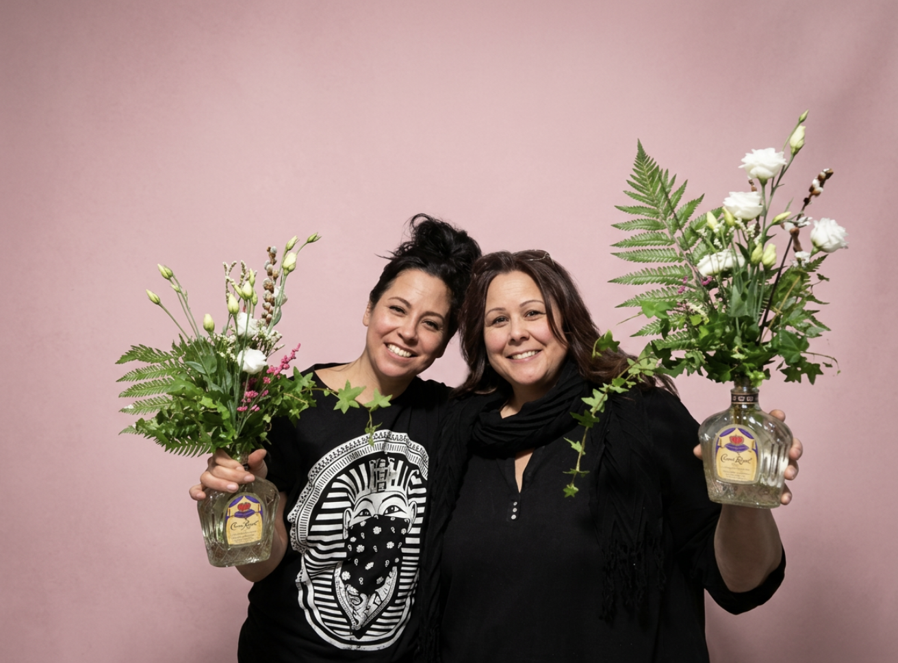 Two women smiling, holding flower arrangements in glass vases, standing in front of a pink background.