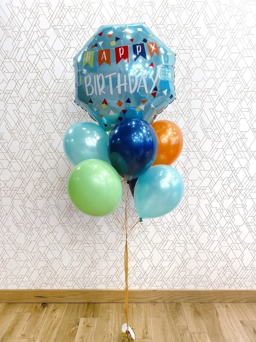 Colorful helium balloons, including a large blue balloon with 'HAPPY BIRTHDAY' written on it, arranged together for a celebration, set against a patterned wall on a wooden floor.