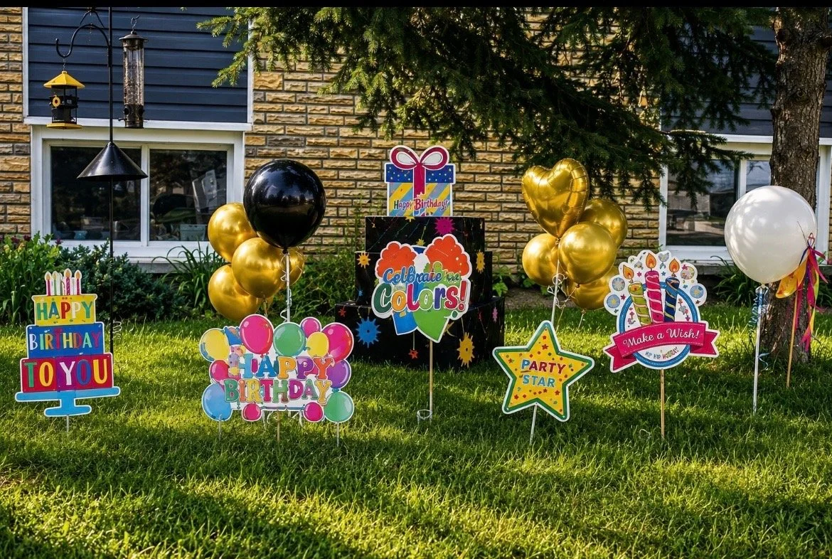 Colorful birthday decorations in a front yard, including balloons and signs with messages like "Happy Birthday to You," "Celebrate the Colors," "Party Star," and "Make a Wish!" set against a brick house and green grass.