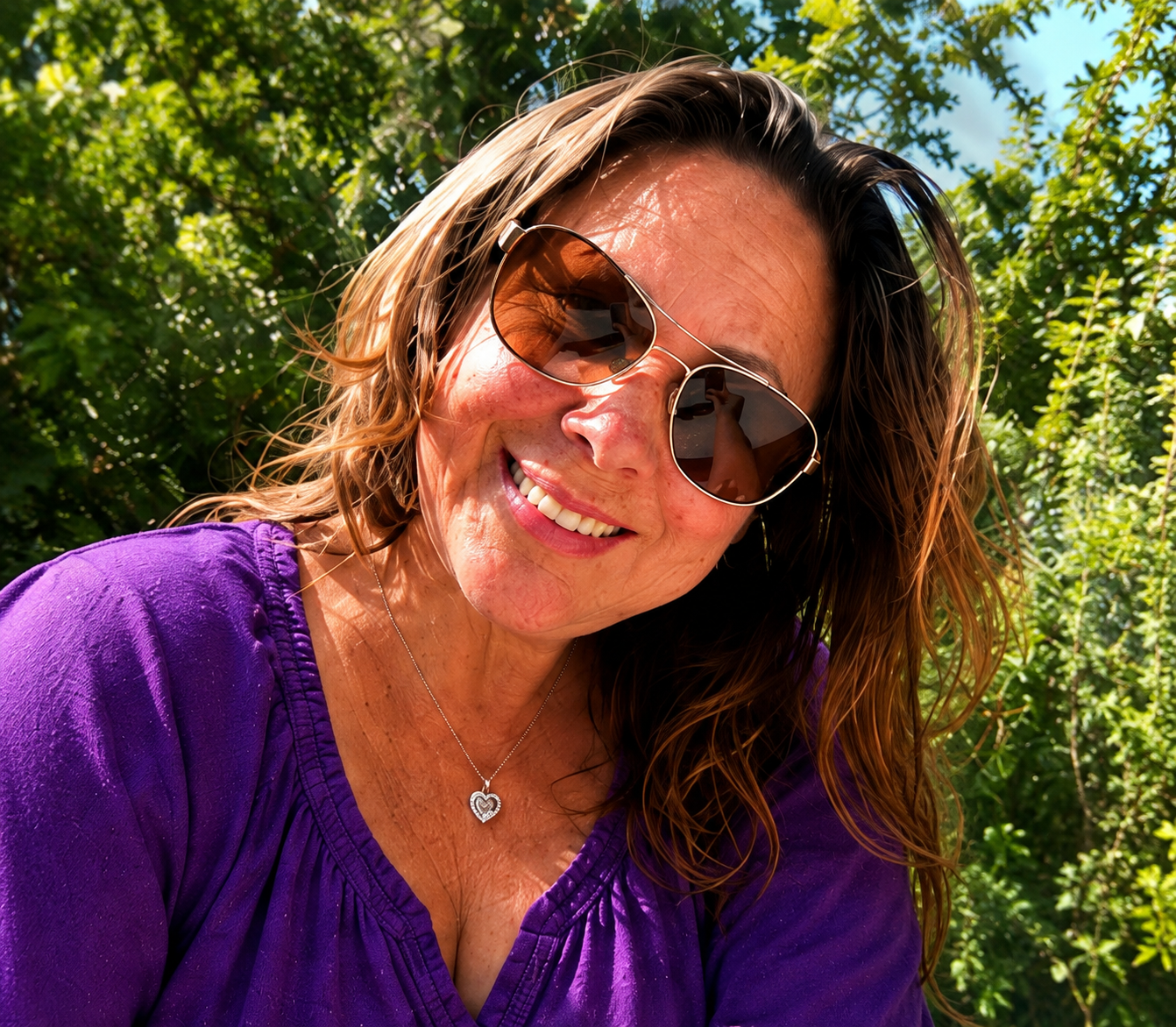 A woman with wavy brown hair wearing sunglasses and a purple top outdoors, smiling with green trees in the background.