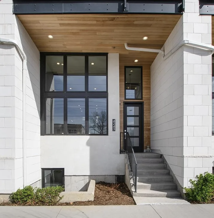 Modern front entrance of a house with a black door, large windows, and steps leading up to the porch. There are small bushes at the base and a wooden ceiling under the overhang.