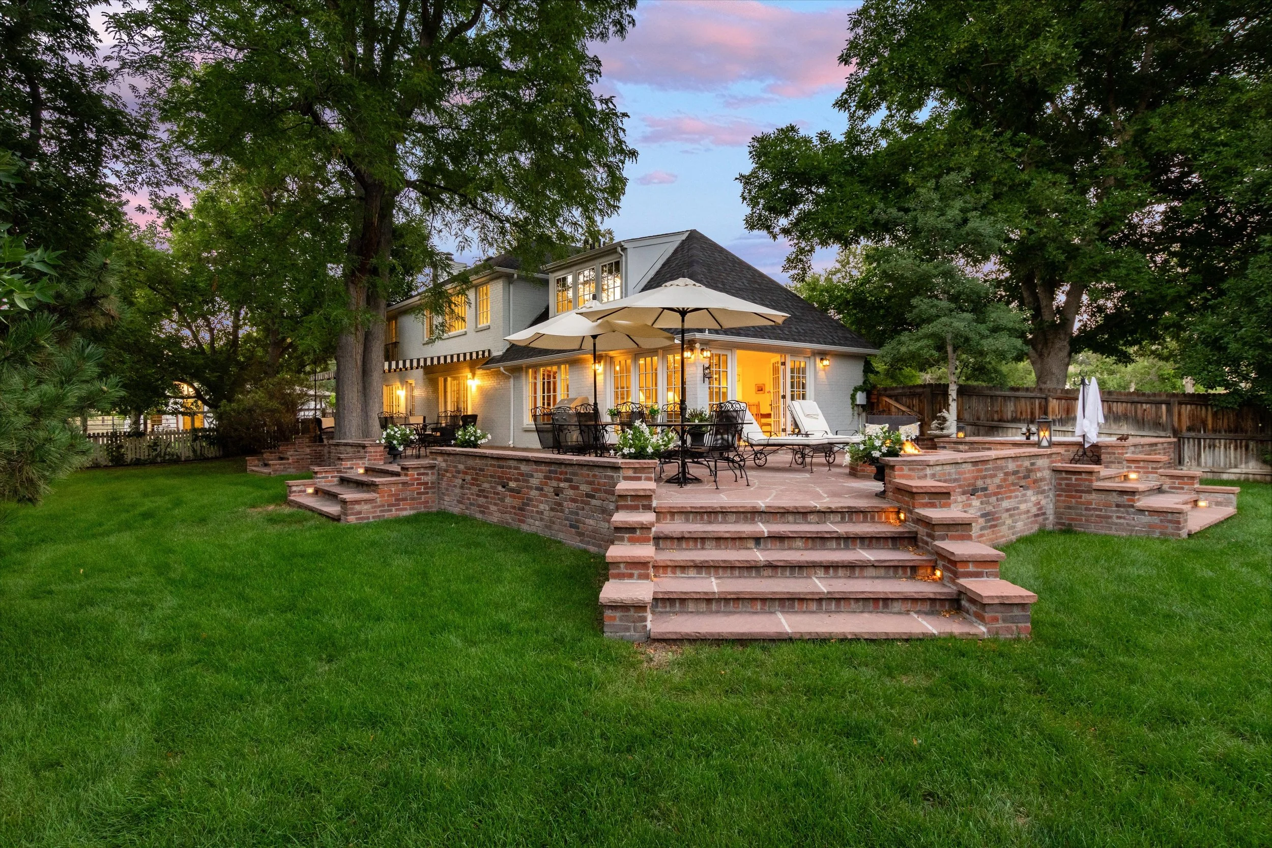 A backyard patio area at dusk with outdoor furniture, umbrellas, potted plants, and string lights, surrounded by trees and a fenced yard. A two-story house with lit windows is in the background.