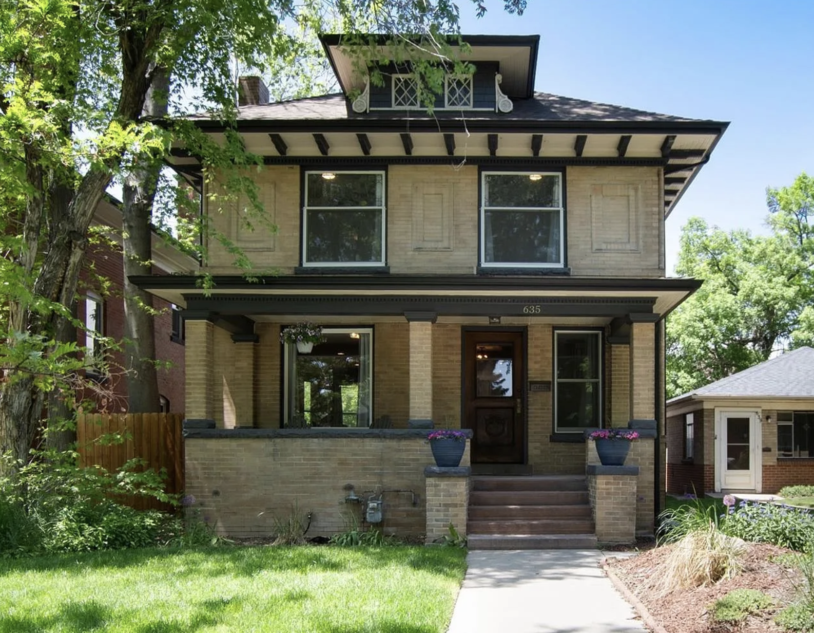 A three-story house with a brick exterior, black trim, and a front porch decorated with potted flowers, leading to a walkway and a well-manicured lawn, surrounded by trees.