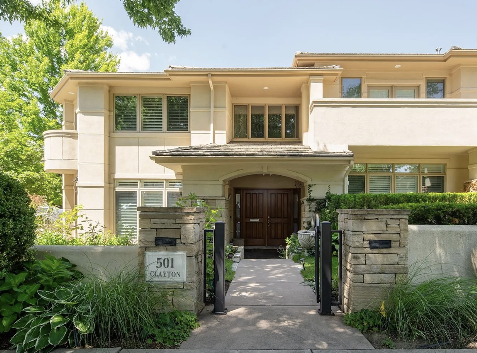 Front view of a multi-story beige house with a landscaped garden and stone fence, featuring a wooden front door and several large windows.
