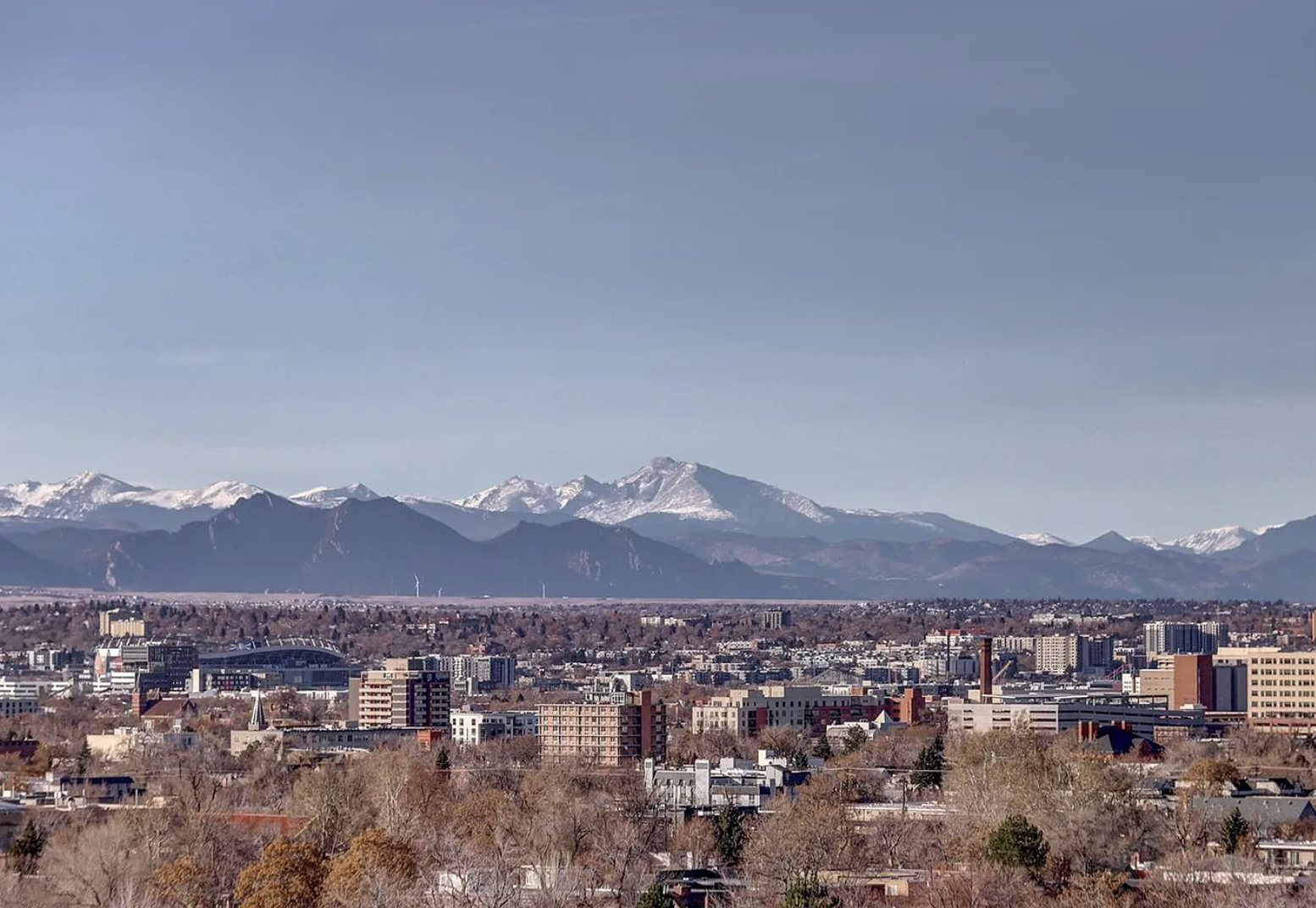 A cityscape with numerous buildings and houses in the foreground, and snow-capped mountains in the background under a clear sky.