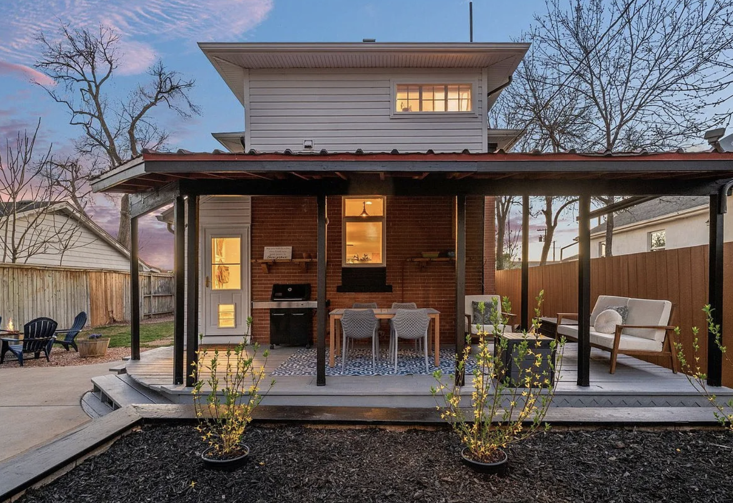 Backyard patio of a two-story house with a covered porch, outdoor furniture, and a grill, during dusk with trees and neighboring houses visible.