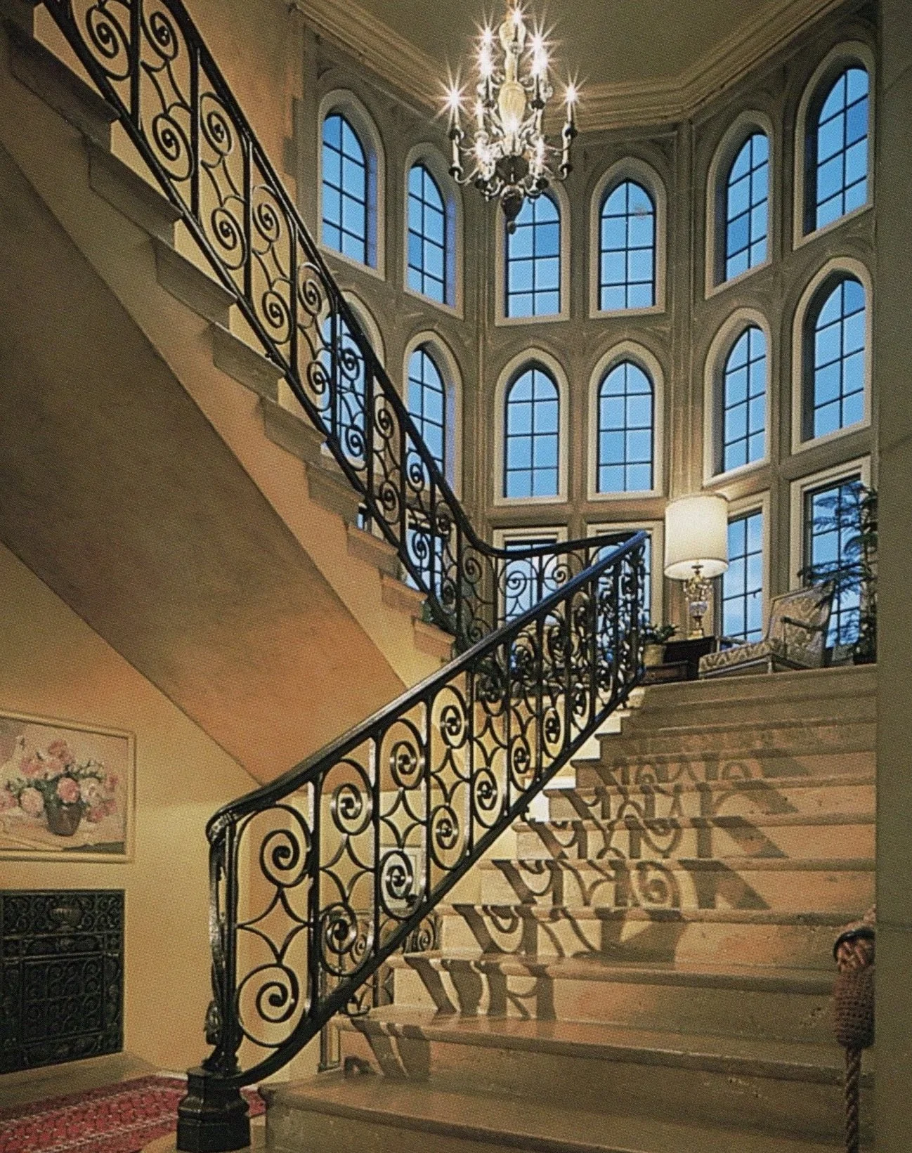Elegant staircase with ornate black wrought iron railing, leading up to a second level with large arched windows, a chandelier hanging from the ceiling, and a sitting area with a lamp and chair near the windows.